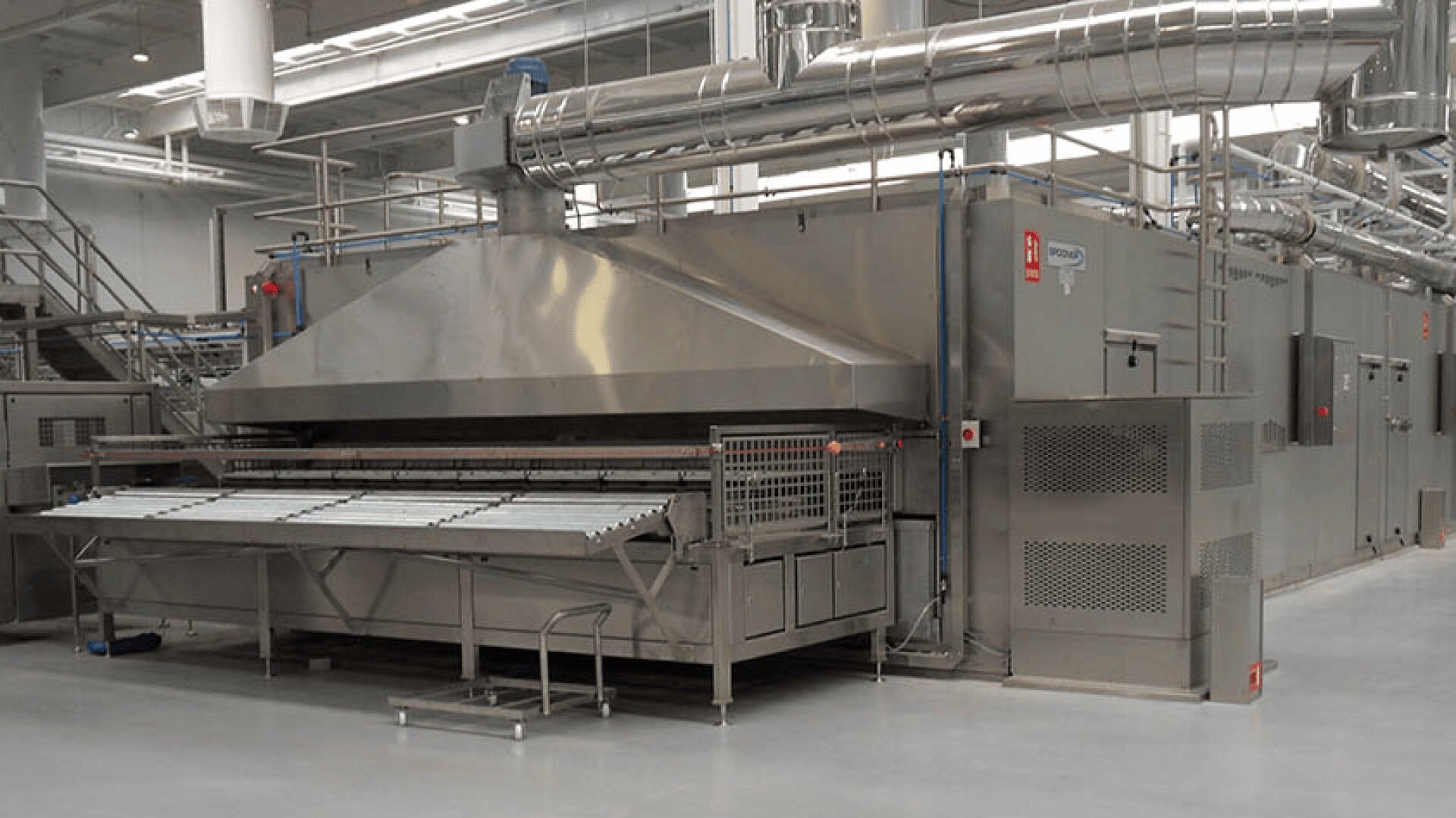 A large industrial bread oven made of stainless steel with conveyor belts and ventilation pipes in a spacious factory setting. The area is clean and brightly lit.