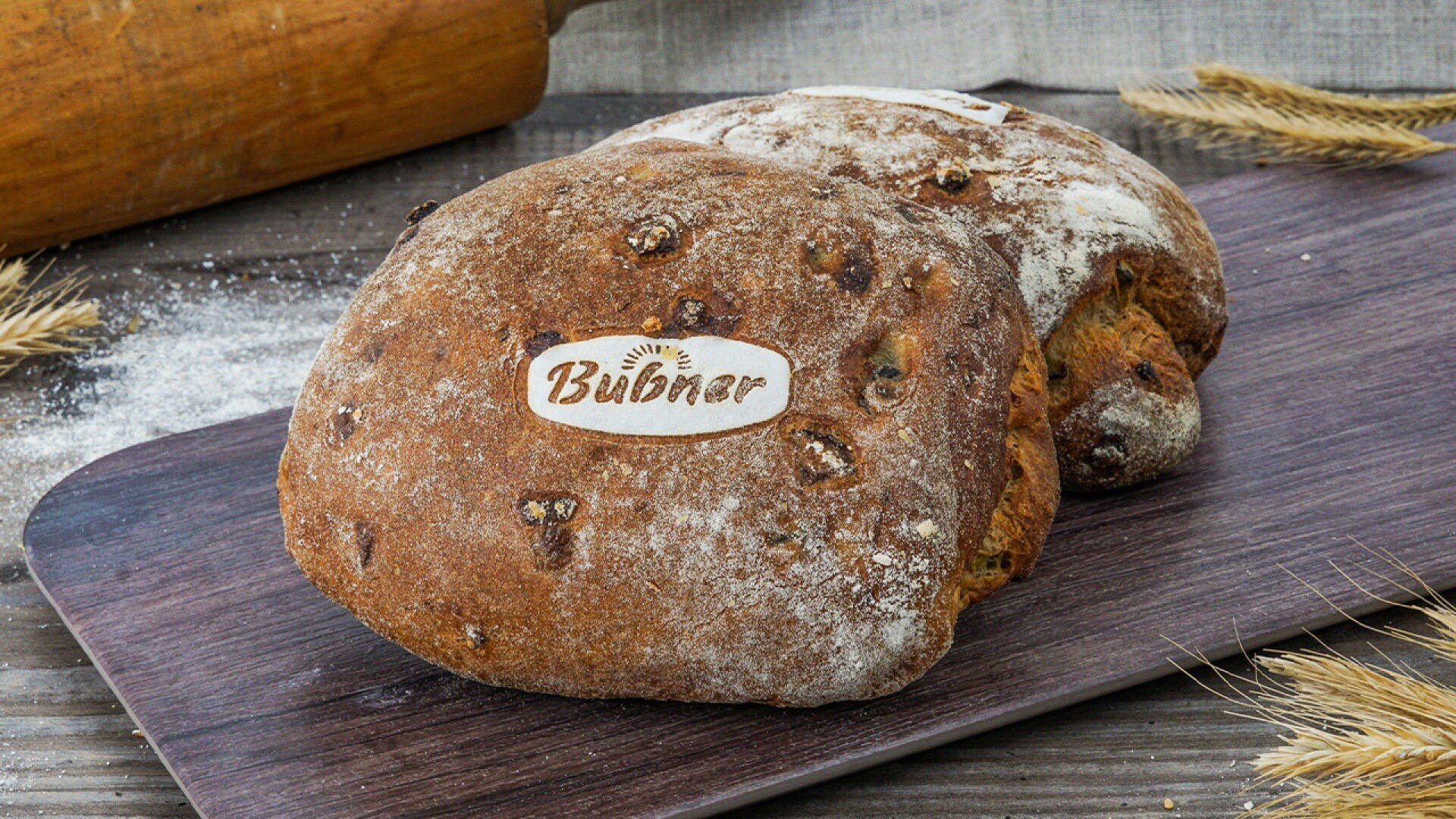 Zwei rustikale Brote mit der Aufschrift „Bubner“ liegen auf einem Holzbrett, umgeben von Weizenhalmen und Mehl, im Hintergrund ist ein Nudelholz zu sehen.