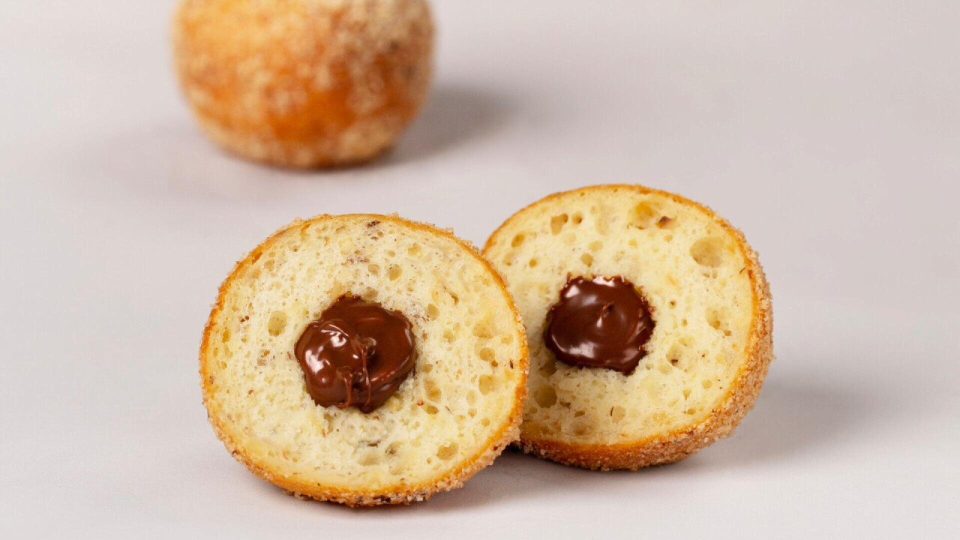 A round donut cut in half showing a chocolate filling inside, with another whole donut in the background on a light surface.