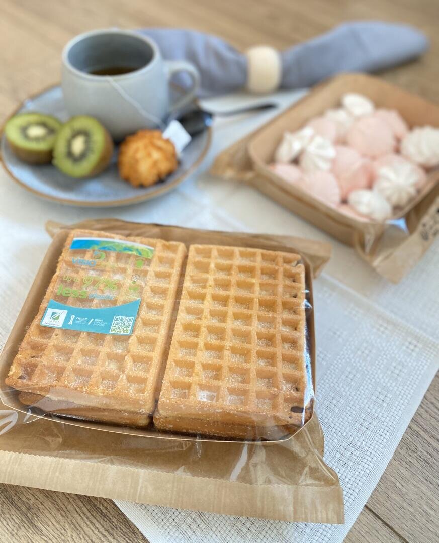 A breakfast scene with packaged waffles in the foreground, a plate with sliced kiwi, a pastry, and a cup of coffee in the background, and a tray of pink and white meringues on a wooden table.