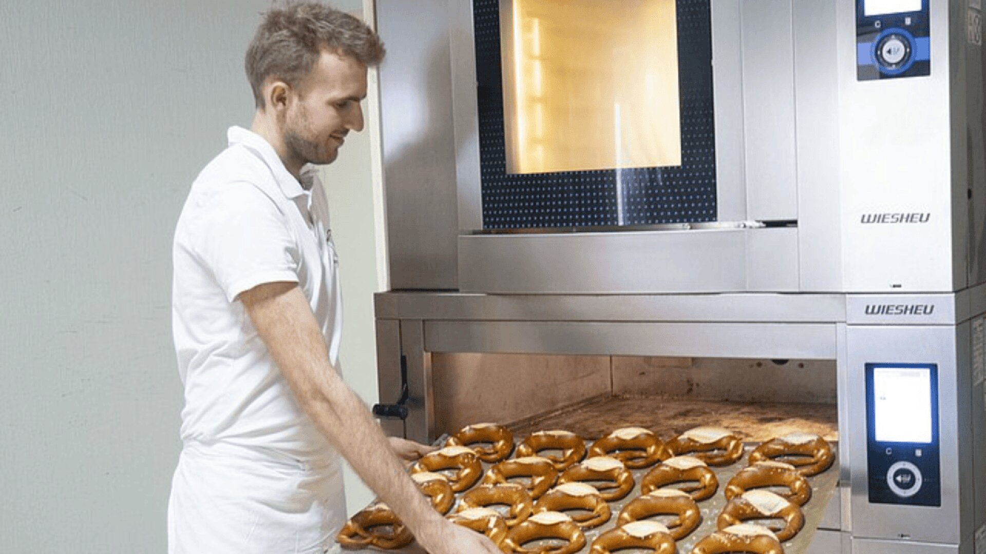 A baker wearing white clothes takes a tray of freshly baked pretzels out of a large industrial oven. The pretzels are golden brown and arranged neatly on the tray.