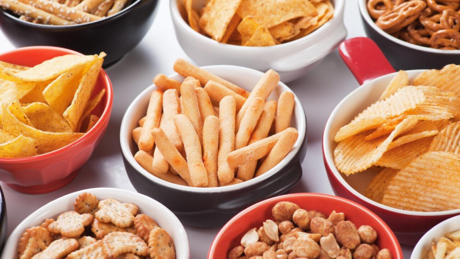 Various bowls filled with different snacks, including tortilla chips, pretzels, potato chips, breadsticks, crackers, and mixed nuts, arranged closely together on a white surface.