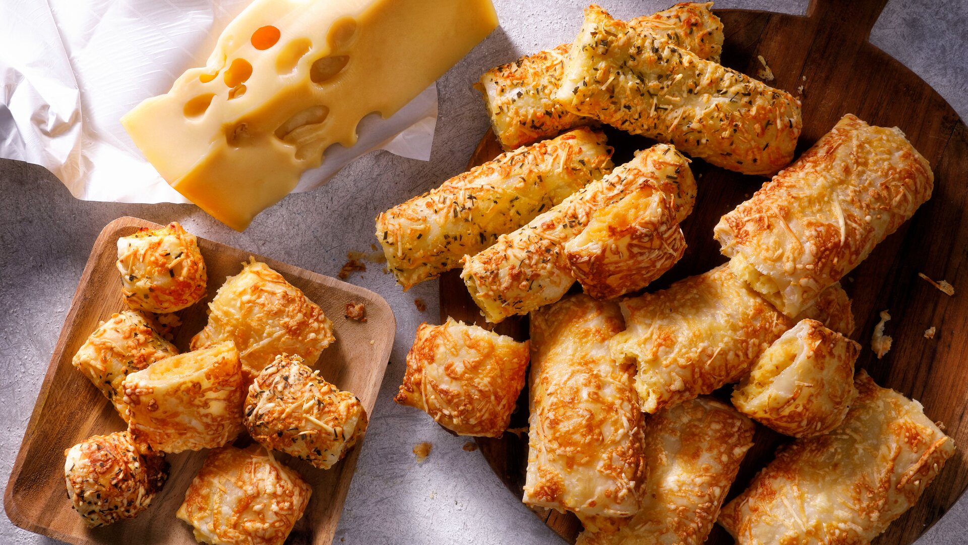 Assorted golden, flaky cheese pastries on wooden boards, next to a block of Swiss cheese with holes, all displayed on a light gray surface.