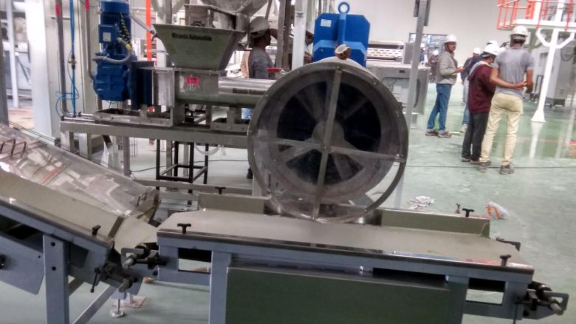 Industrial food processing equipment in a factory setting, with metal machinery, conveyor belts, and several workers wearing safety helmets and uniforms standing in the background.