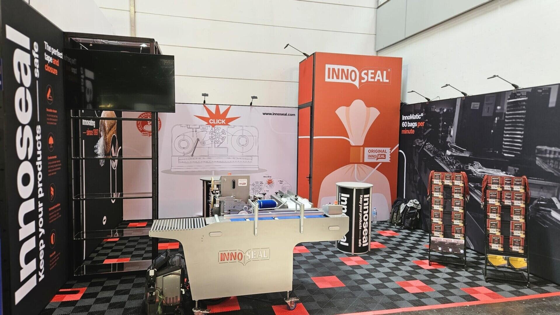 Exhibition booth for InnoSeal featuring a sealing machine in the center, branded banners, product displays, and a TV screen, set up on a red-and-black checkered floor in a well-lit space.