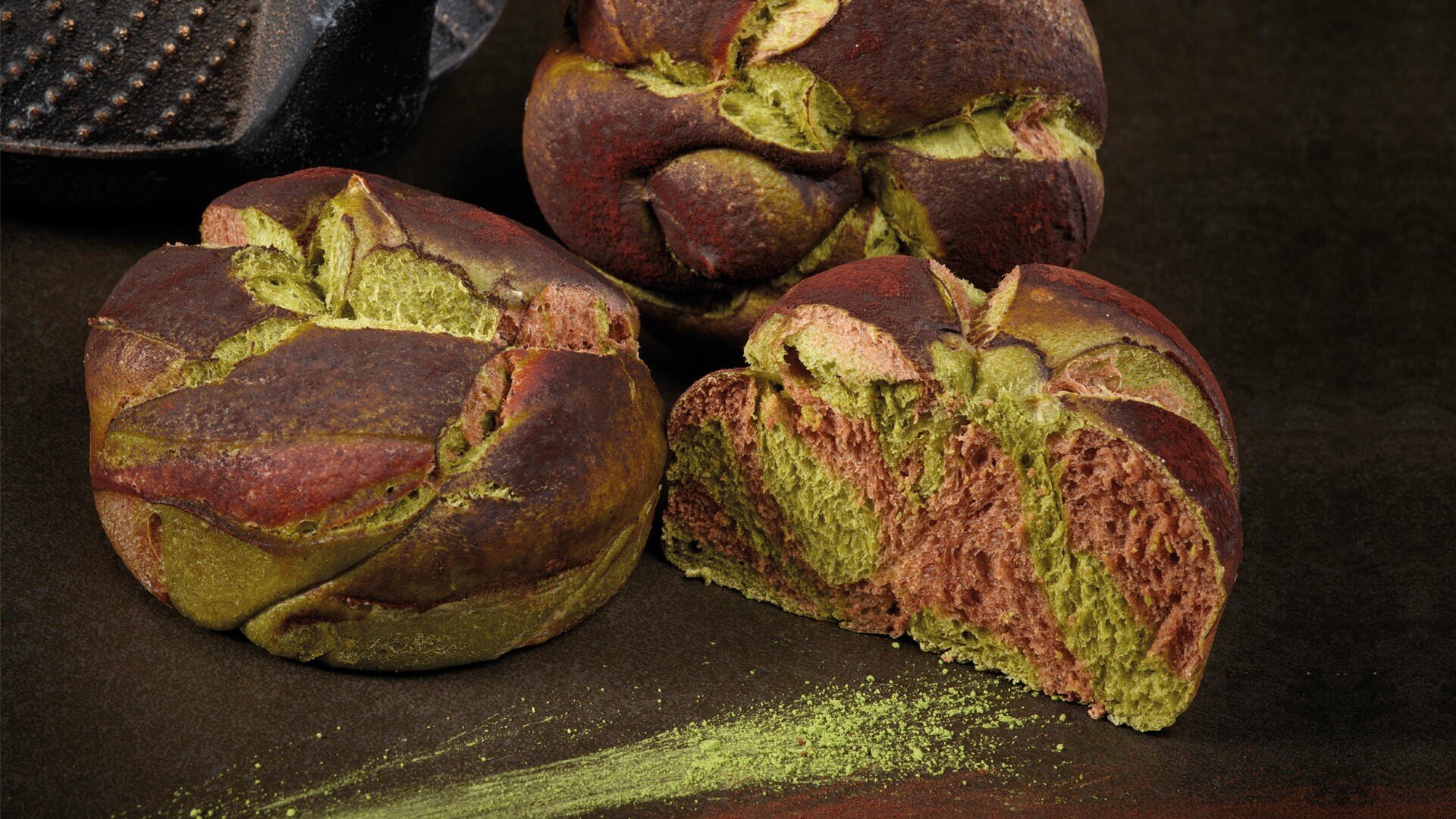 Three round loaves of marbled bread with green and brown swirls, with one loaf cut open to reveal its colorful interior. Green matcha powder is sprinkled on the dark surface next to it.