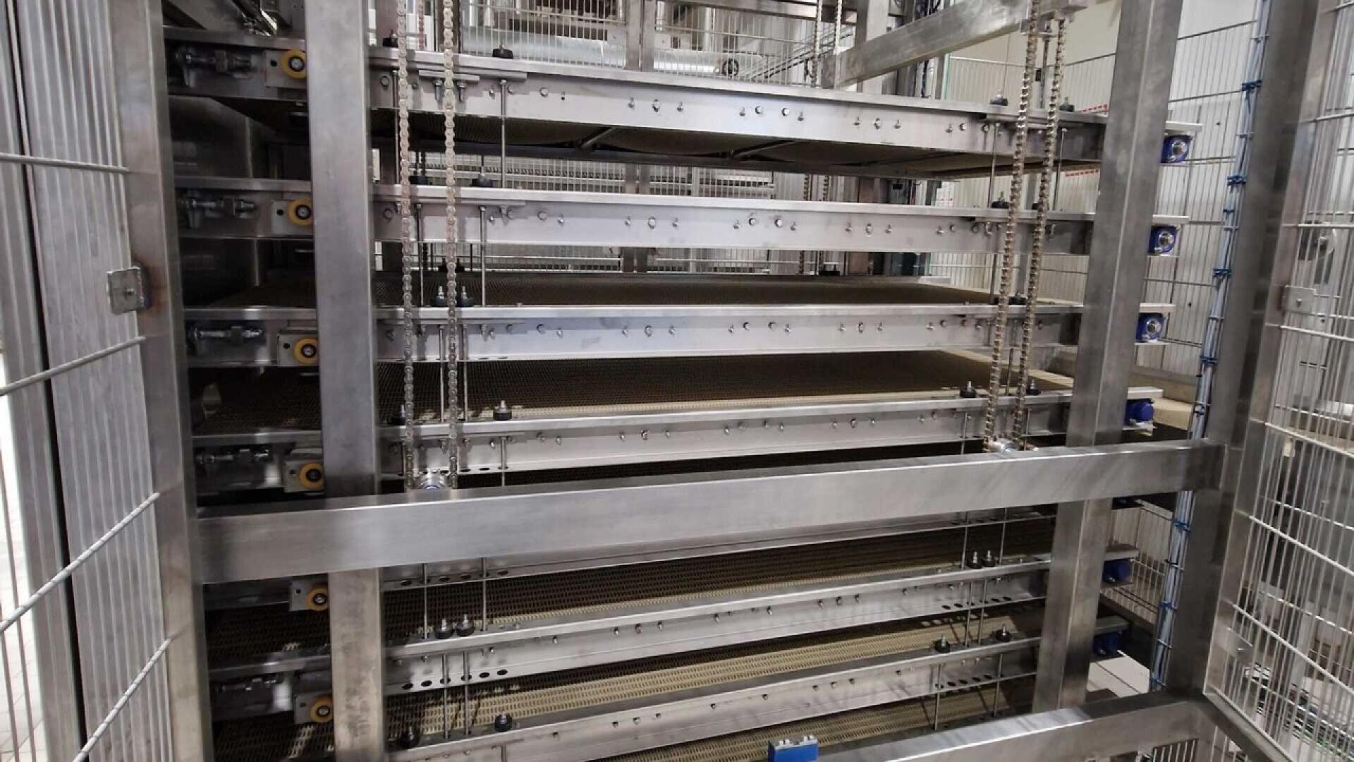 A close-up view of a multi-tiered metal structure, possibly an industrial shelving or conveyor system, with horizontal trays or belts and vertical metal supports inside a fenced area.