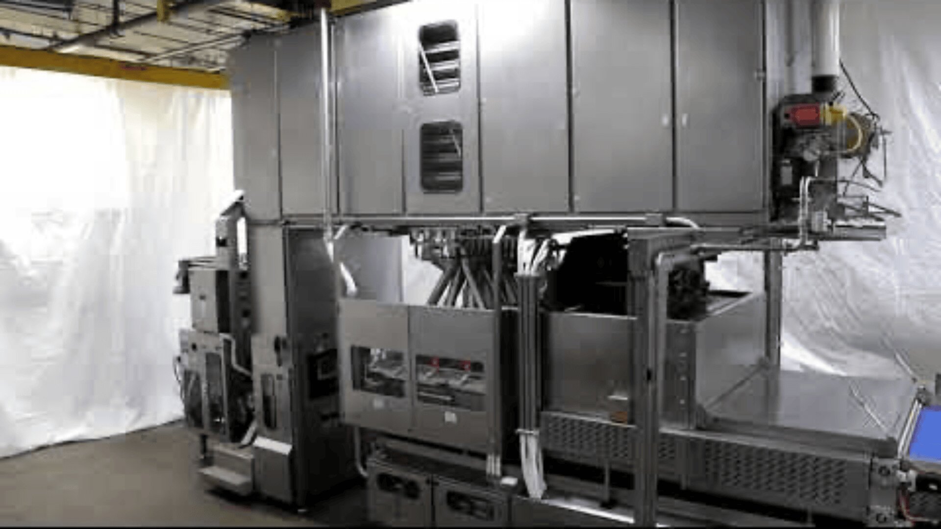 Large stainless steel industrial machine with vents, control panels and various pipes, standing in an interior space in front of a white curtain on a concrete floor.