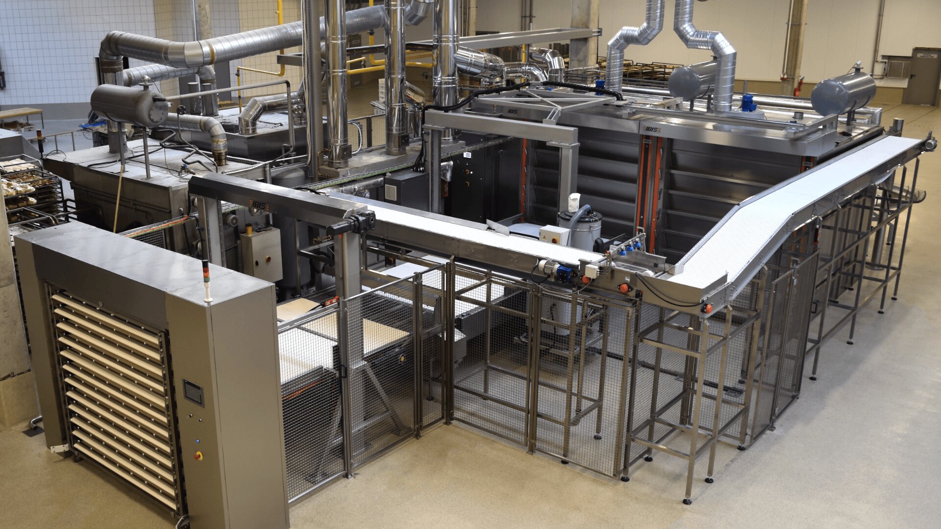 A modern industrial food processing machine with conveyor belts, metal structures, and ventilation pipes in a clean factory setting. Trays are visible in a rack on the left side of the equipment.