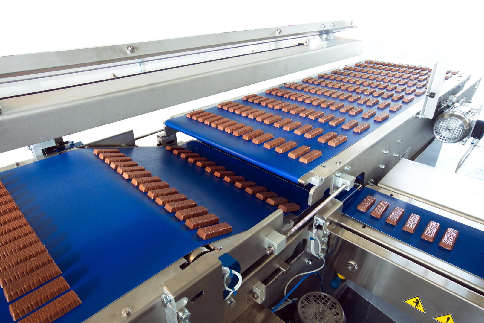 Rows of chocolate bars move along blue conveyor belts in a modern factory setting, with metal machinery surrounding the belts as the chocolates are processed or packaged.