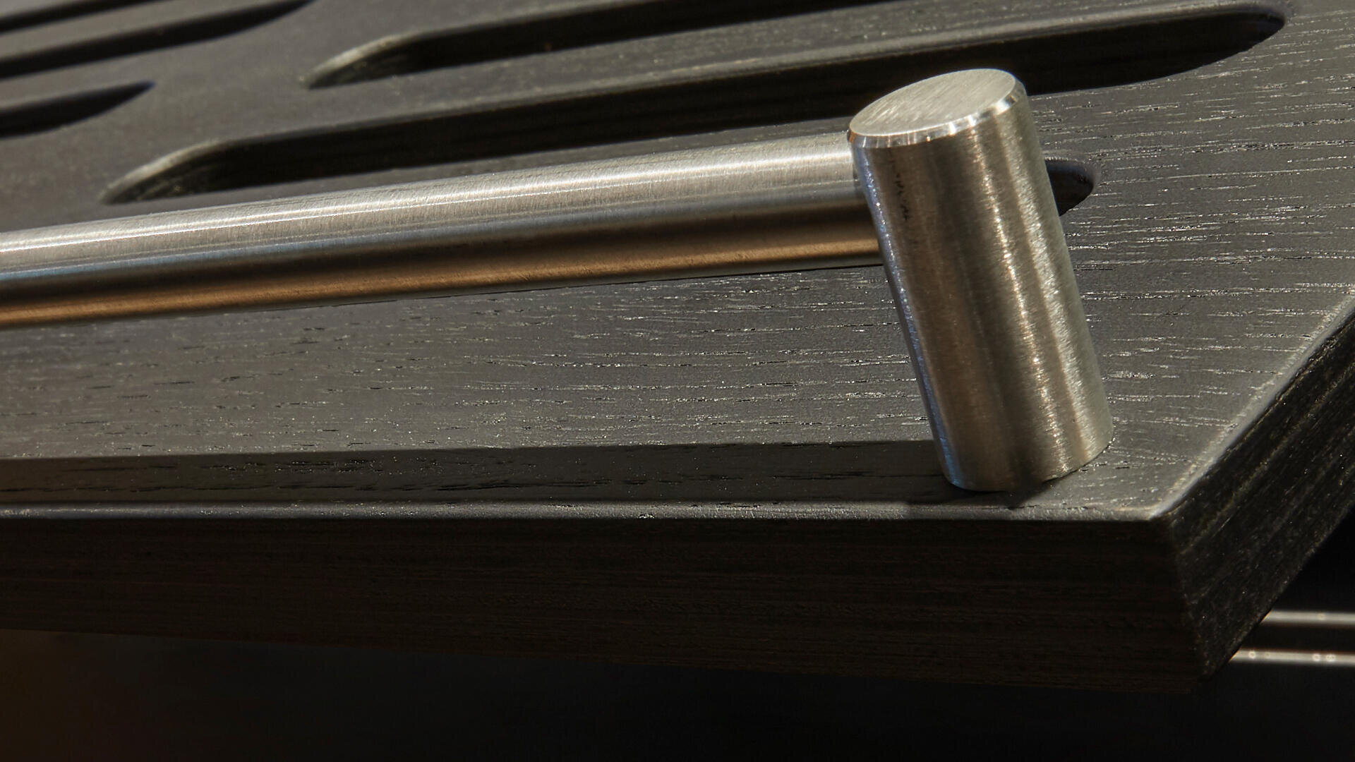 Nahaufnahme eines zylindrischen Metallgriffs, der an einer dunklen, strukturierten Holzoberfläche mit länglichen Schlitzen oder Rillen befestigt ist.