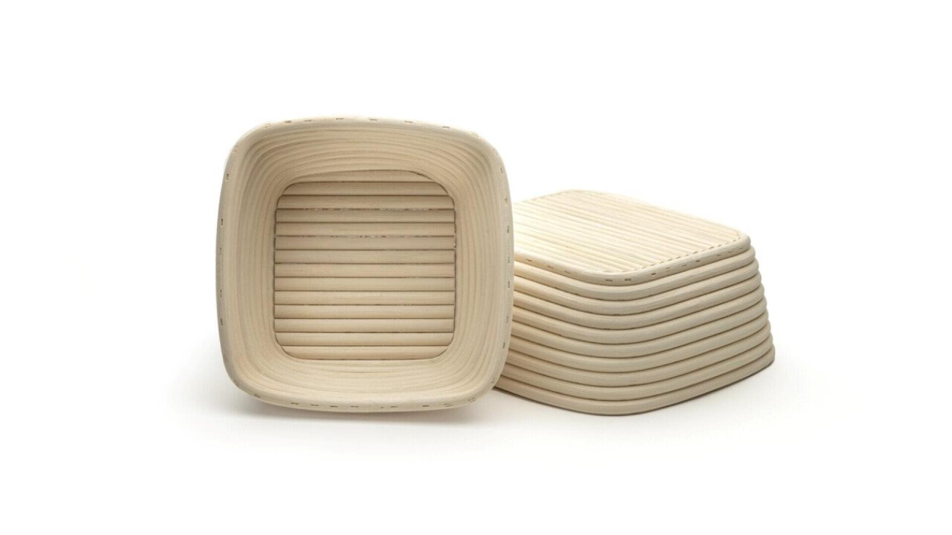 Two square, light-colored, ribbed proofing baskets made of woven material are shown. One basket is standing upright, while a stack of similar baskets is placed on its side behind it. The background is white.