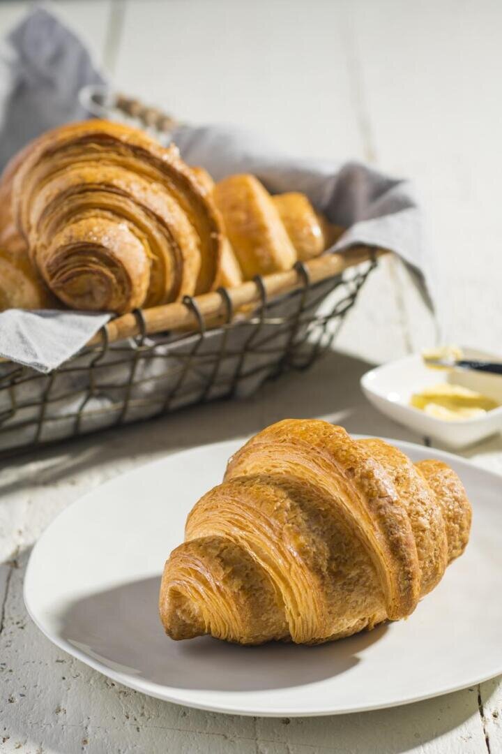 Ein goldenes, flockiges Croissant liegt auf einem weißen Teller im Vordergrund, während ein Korb mit weiteren Croissants und eine Schale mit Butter im Hintergrund auf einem weißen Holztisch stehen.
