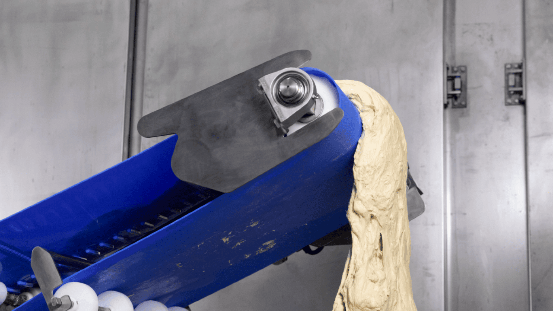 Ein Förderband mit blauer Grundfläche transportiert eine große Menge hellen Teigs, der in einer industriellen Umgebung von einem metallischen Maschinenteil geformt wird.