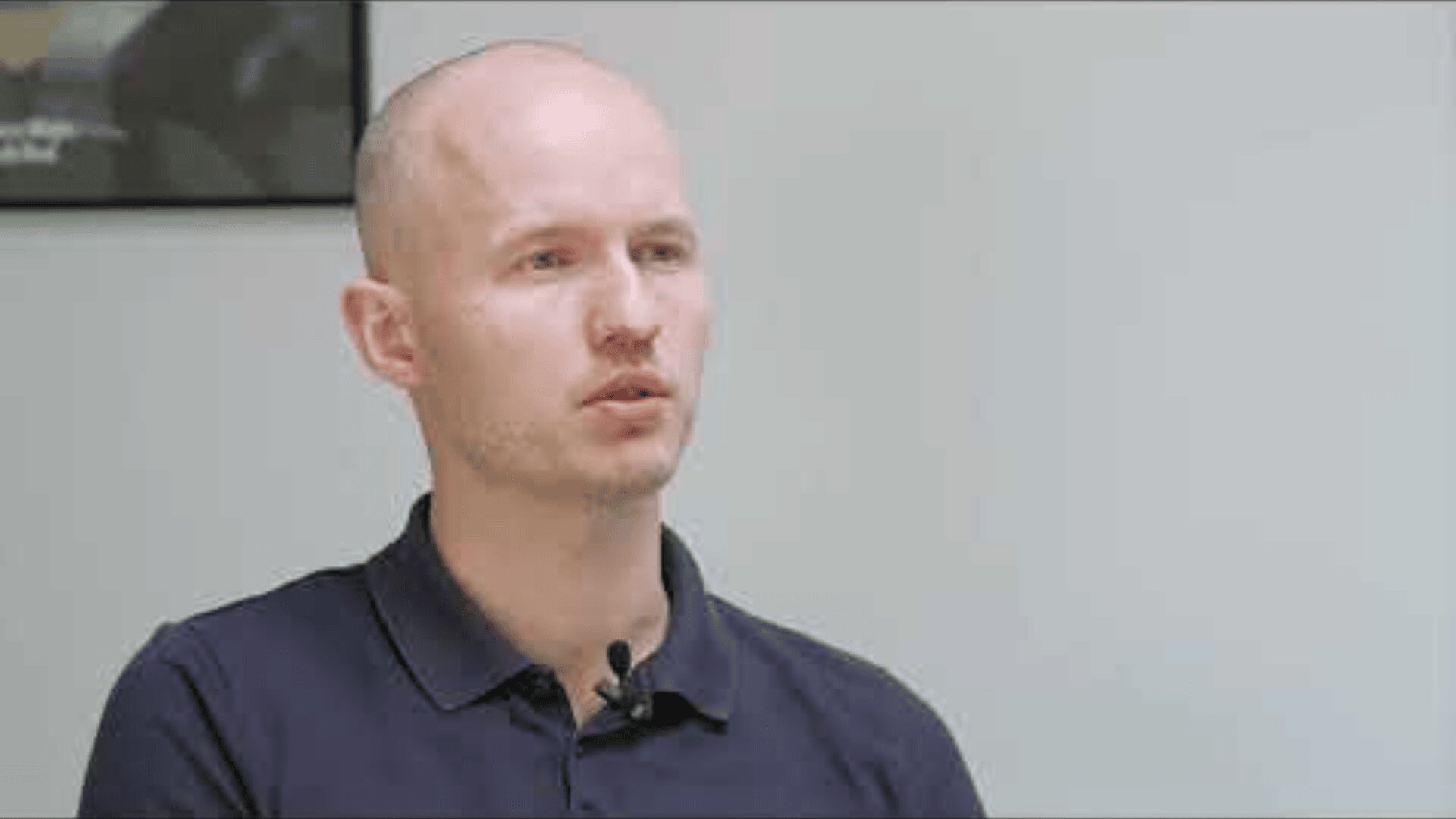 A man with a shaved head wearing a navy blue polo shirt speaks while seated indoors. He has a small microphone clipped to his shirt, and the background is plain and light-colored.