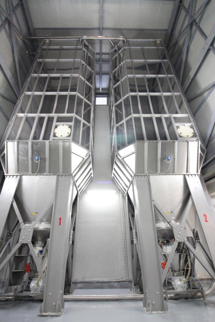 Two large, industrial metal silos with supporting beams and numbered 1 and 2, standing inside a spacious, well-lit facility. The silos have grated platforms and machinery attached at their bases.