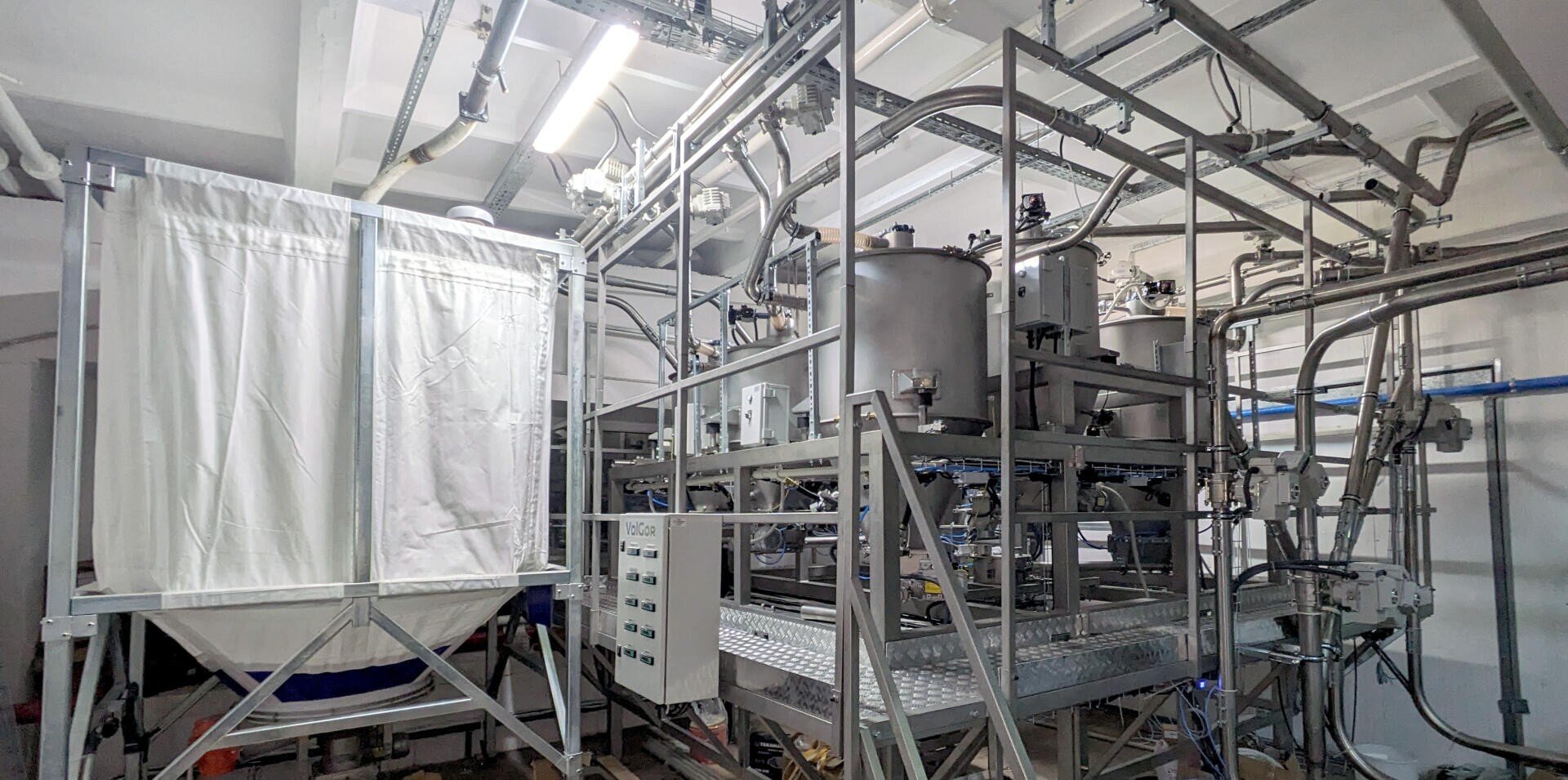 Industrial equipment with metal pipes, tanks, control panels, and a large white fabric container inside a well-lit facility. The machinery appears to be used for processing or manufacturing purposes.