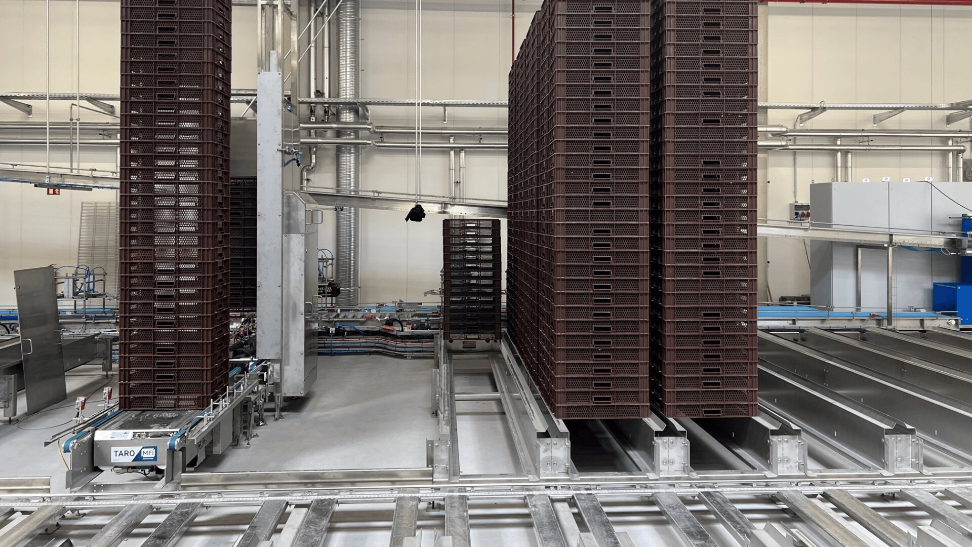 Tall stacks of brown plastic crates are positioned in rows inside an industrial facility, with metal conveyor tracks and various machinery visible throughout the clean, spacious area.