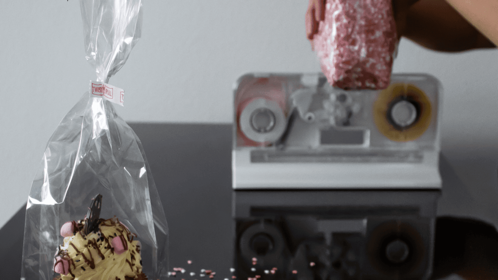 Eine Person wickelt ein dekoriertes Törtchen in durchsichtiges Plastik ein und benutzt dazu einen Klebebandabroller im Hintergrund. Rosa und weiße Streusel sind auf die glänzende schwarze Oberfläche gestreut.