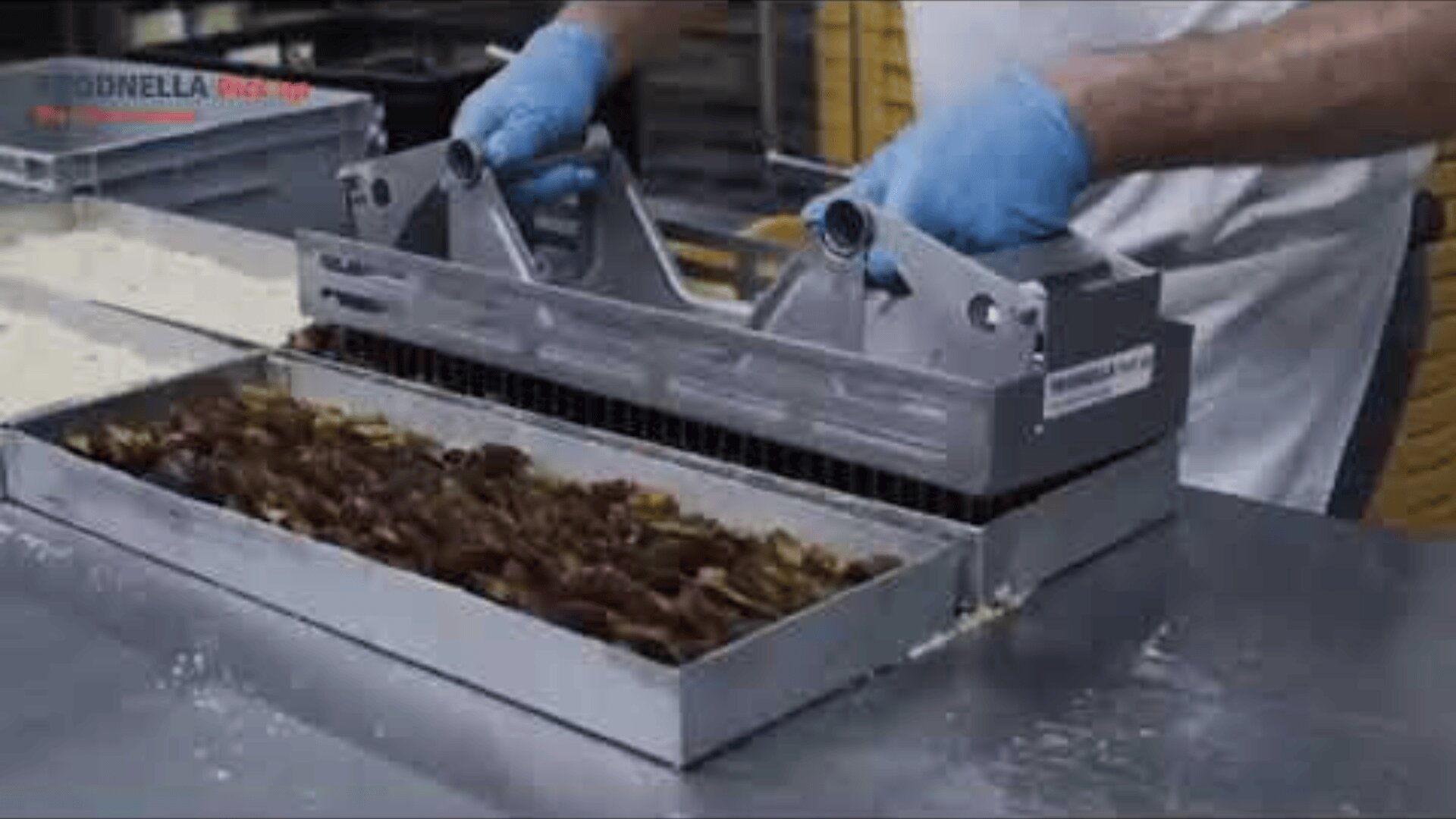 Eine Person mit blauen Handschuhen verteilt oder drückt mit einem Metallwerkzeug eine Schicht Datteln oder eine ähnliche Füllung gleichmäßig in eine rechteckige Metallschale auf einer Arbeitsfläche aus Edelstahl.