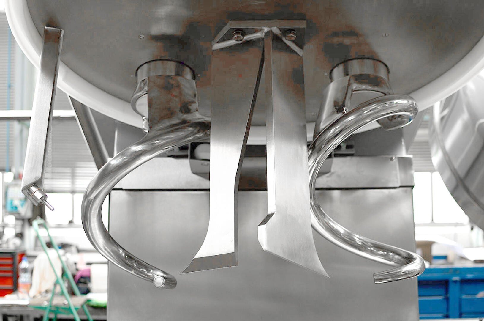 Close-up of a large industrial dough mixer with metal spiral and paddle attachments, set in a commercial kitchen or bakery environment.
