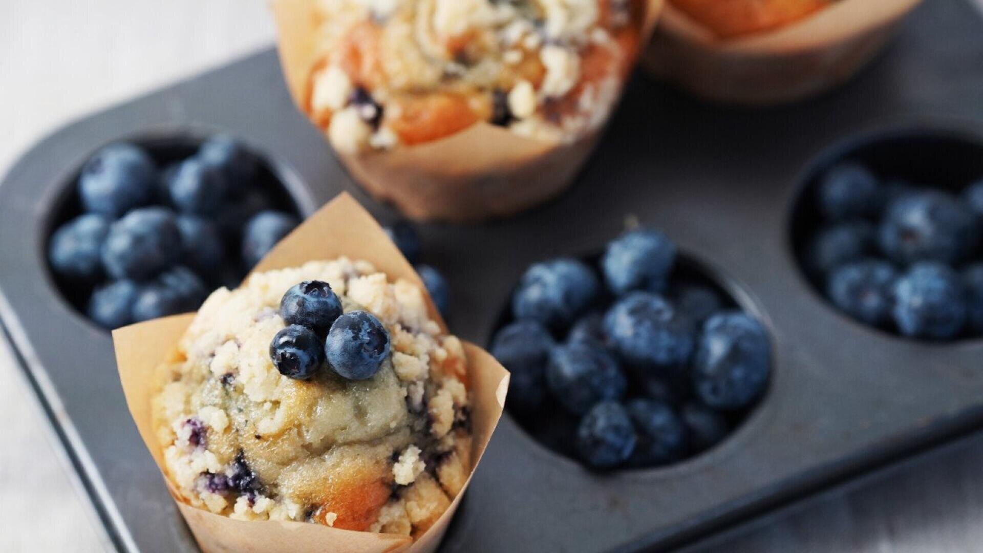A close-up of a blueberry muffin topped with fresh blueberries, placed in a brown paper liner. More blueberries and muffins are visible in a muffin tin in the background.