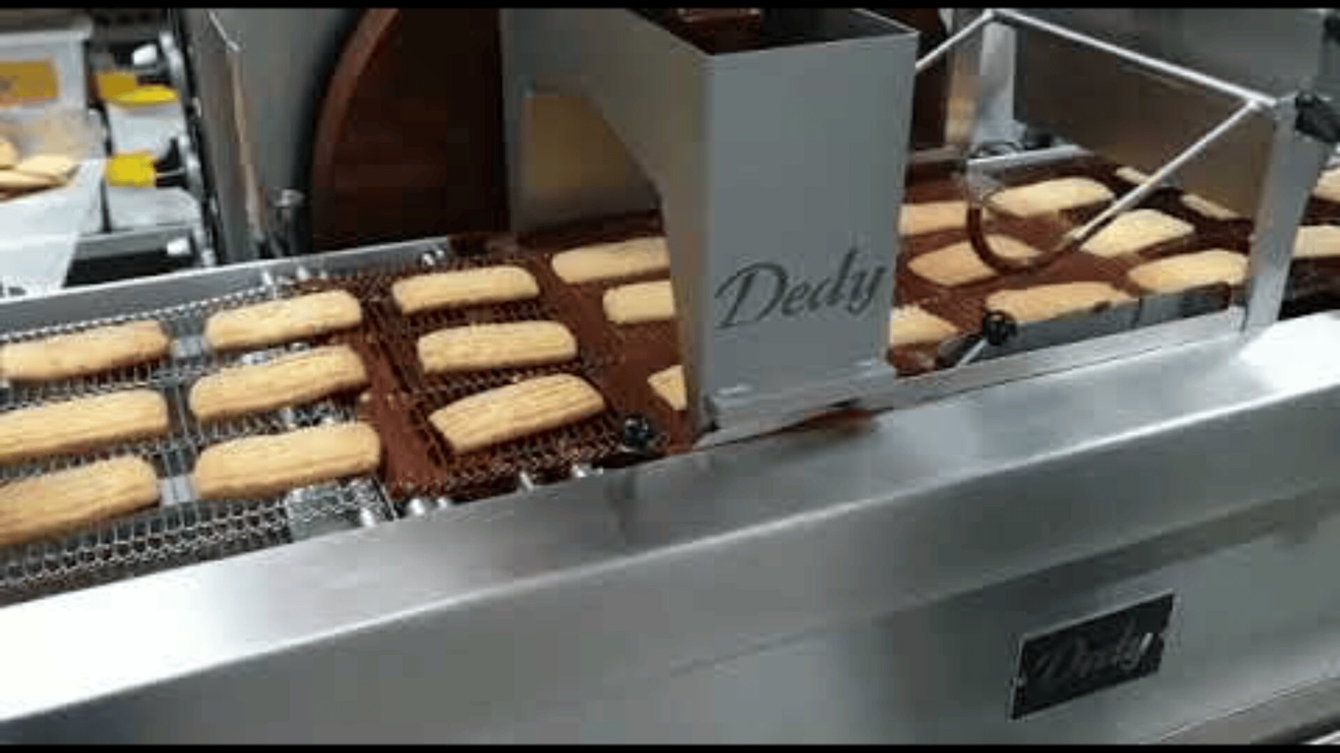 Cookies are moving on a conveyor belt through a metal machine, which appears to be used for enrobing or processing baked goods in a factory setting.