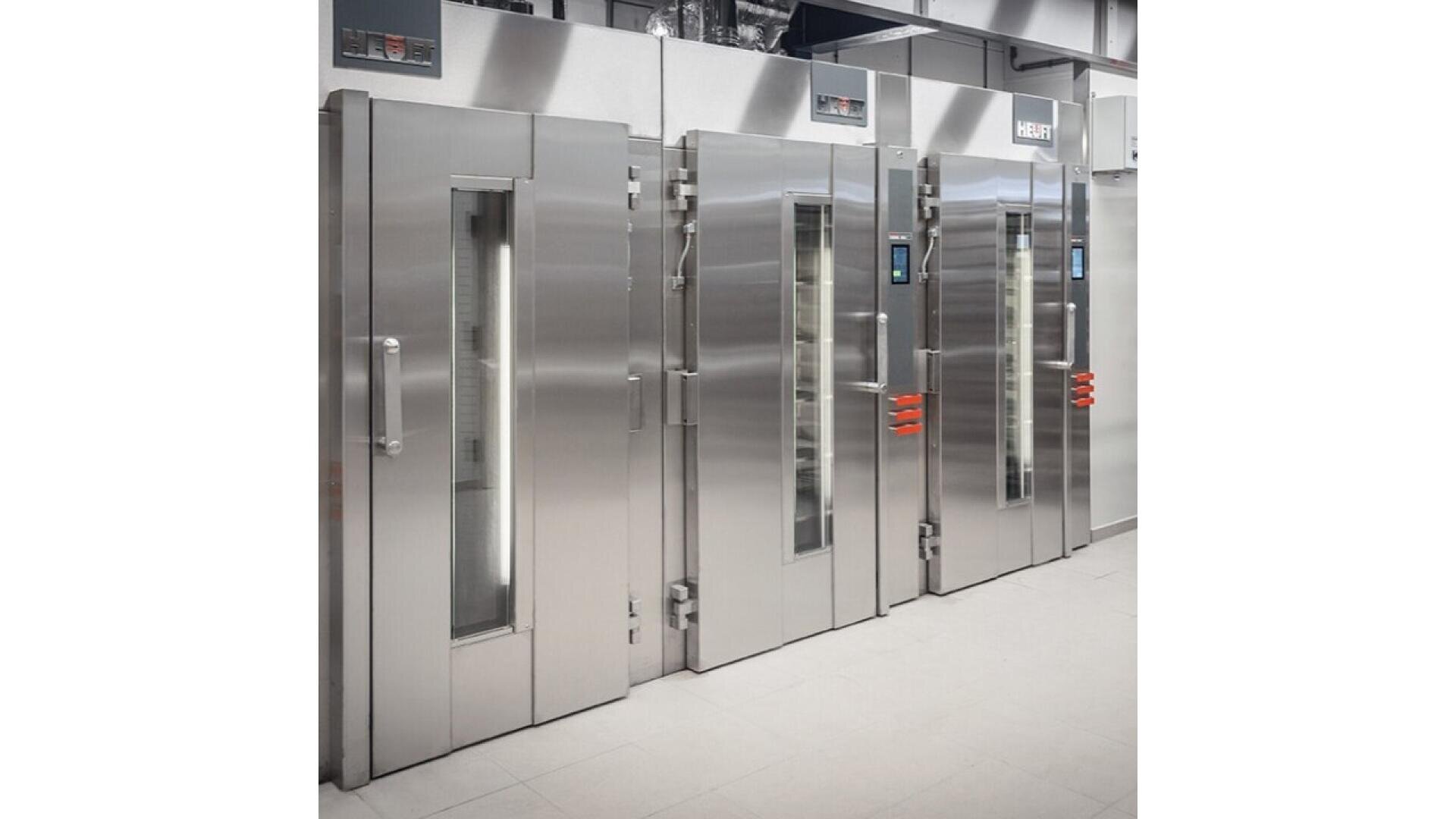 Three large, industrial stainless steel ovens with vertical glass windows and digital controls stand side by side in a clean, modern commercial kitchen.