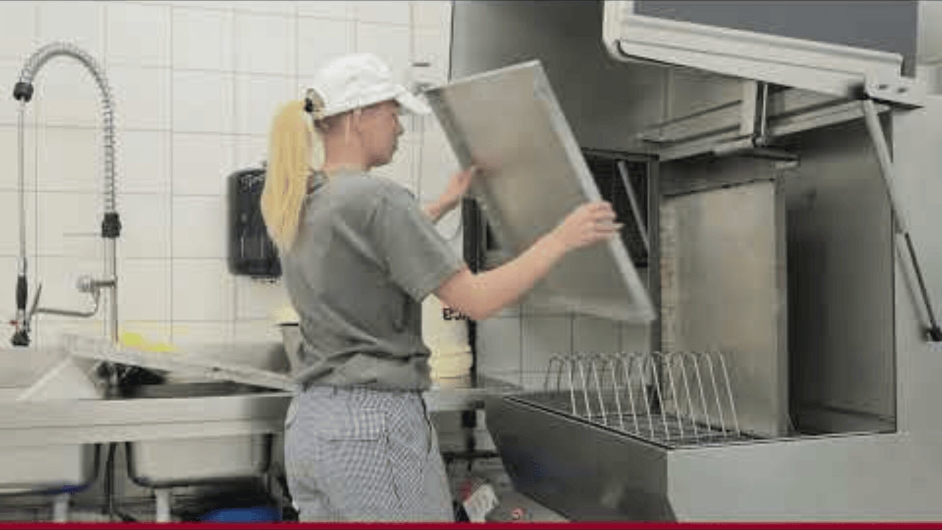 Eine Person in einem grauen Hemd, einer karierten Hose und einer weißen Mütze lädt ein großes Tablett in einen gewerblichen Geschirrspüler in einer gefliesten Küche.