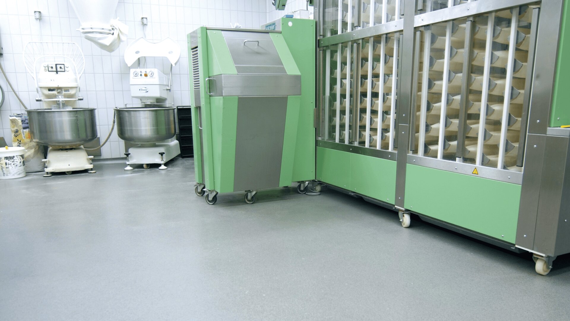 Eine saubere Industrieküche mit grüner Edelstahlbäckereiausrüstung, darunter Knetmaschinen und eine große Teigverarbeitungsmaschine, auf grauem Boden und weiß gekachelten Wänden.