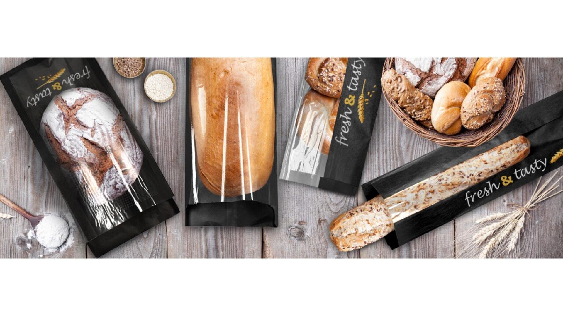 Assorted loaves and rolls of bread in clear, black packaging labeled fresh & tasty, displayed on a rustic wooden surface with grains, flour, and a wicker basket.