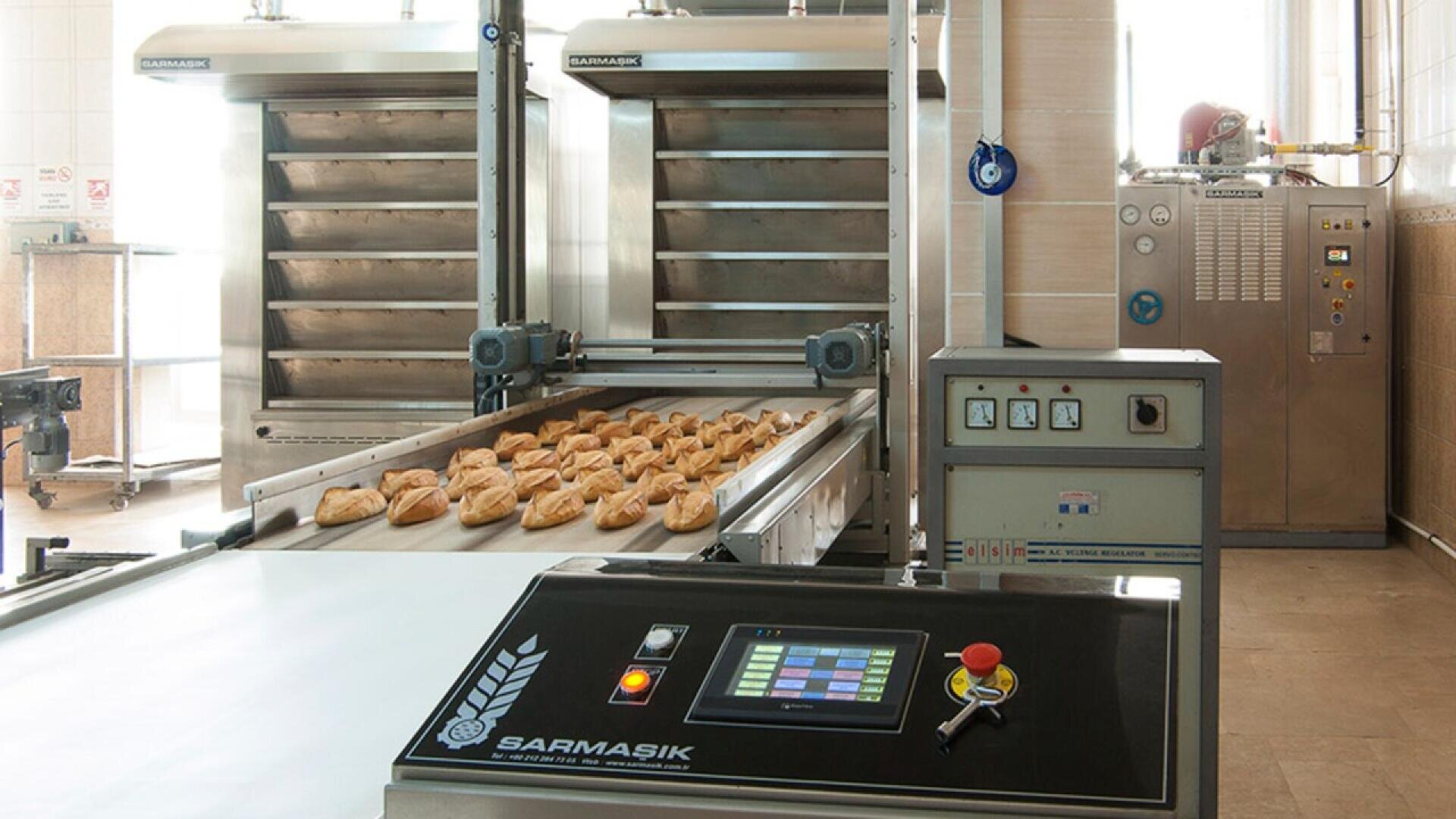 A commercial bakery conveyor system transports freshly baked bread rolls past control panels and large industrial ovens in a modern, clean bakery setting.