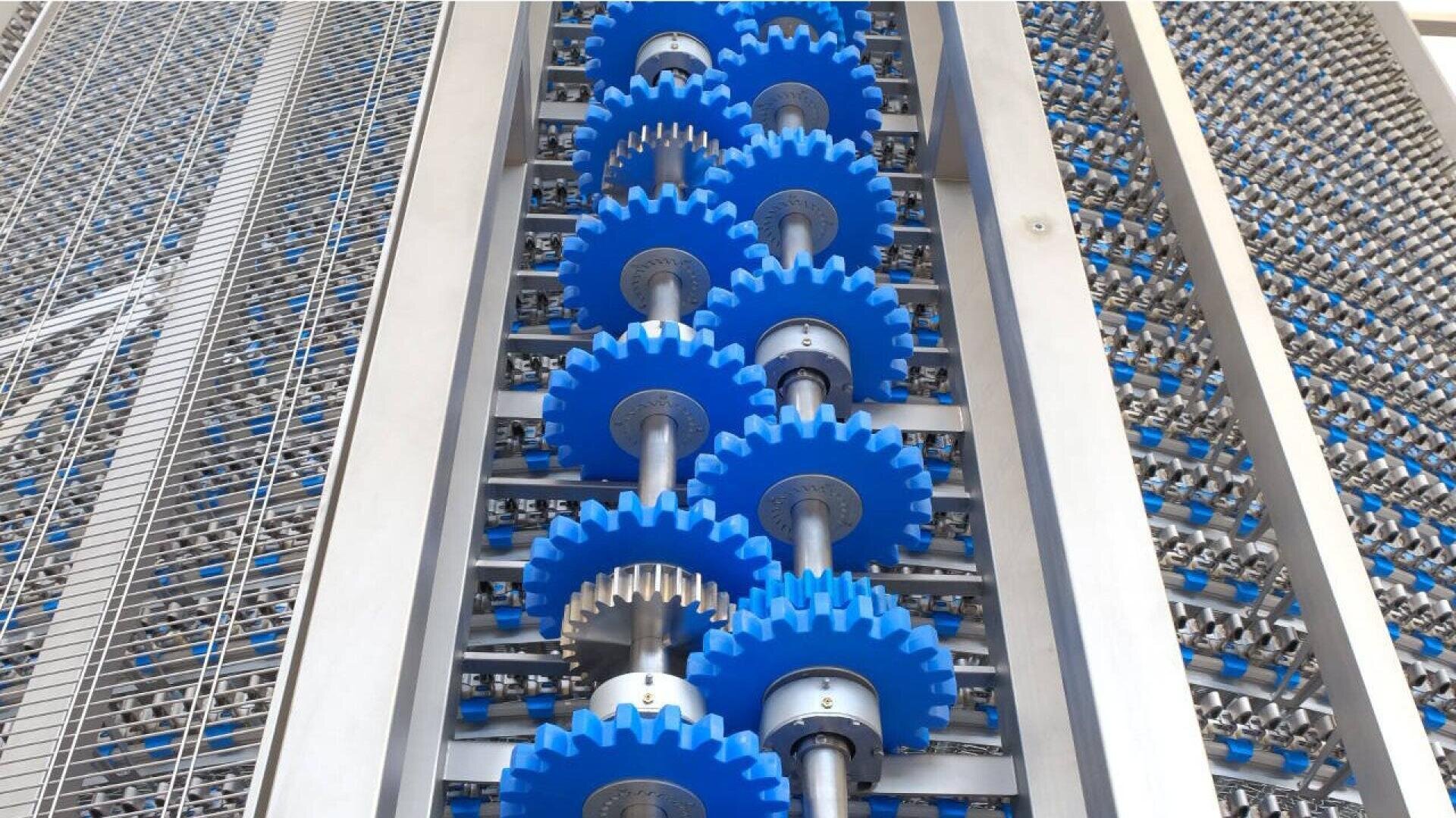 Nahaufnahme von blauen Kunststoffzahnrädern auf vertikalen Metallwellen, Teil einer Industriemaschine, mit Metallgitter und Rollenreihen an den Seiten.