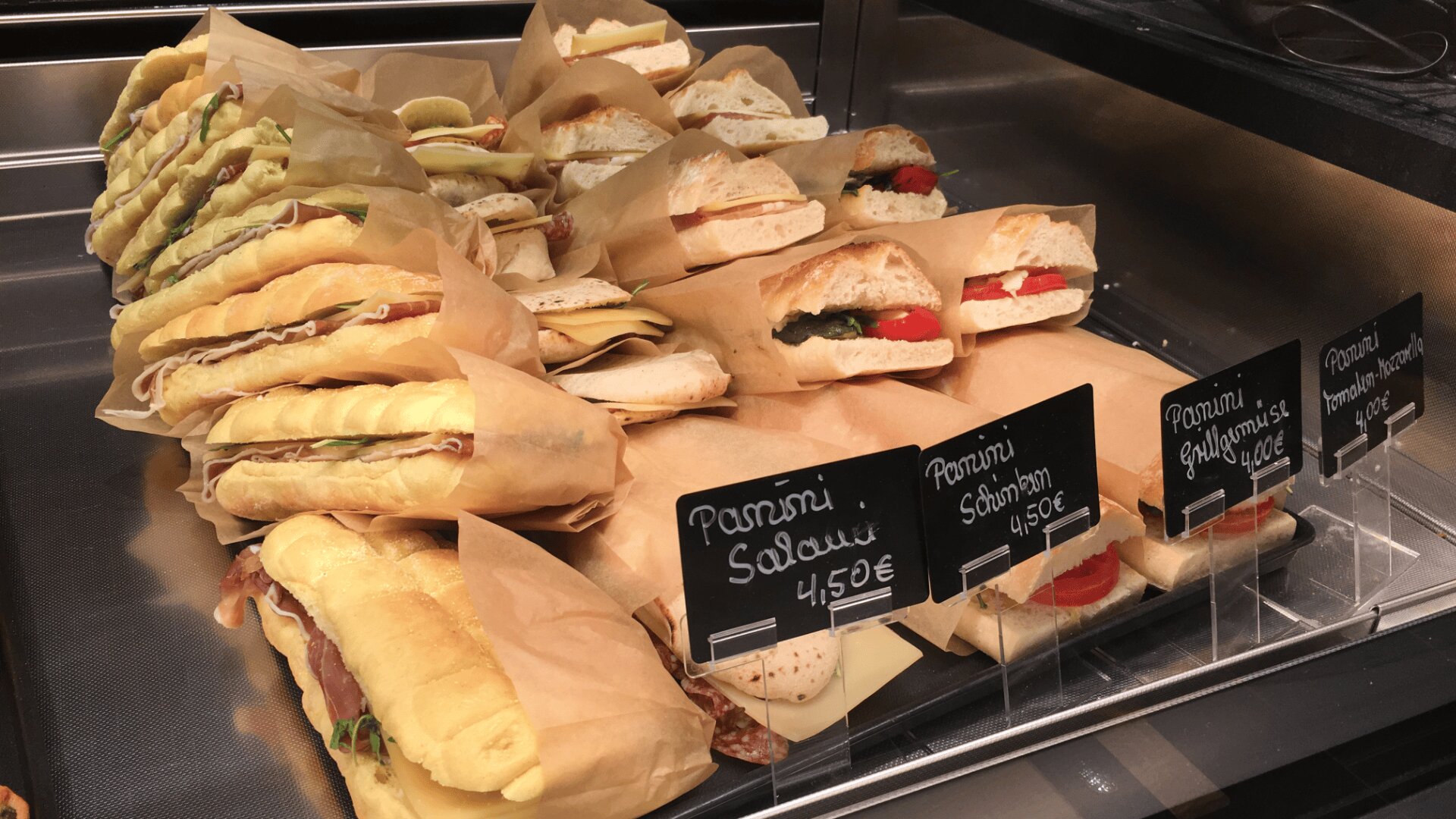 A display case filled with various panini sandwiches wrapped in paper, labeled with signs showing different types and prices, including salami and vegetables.
