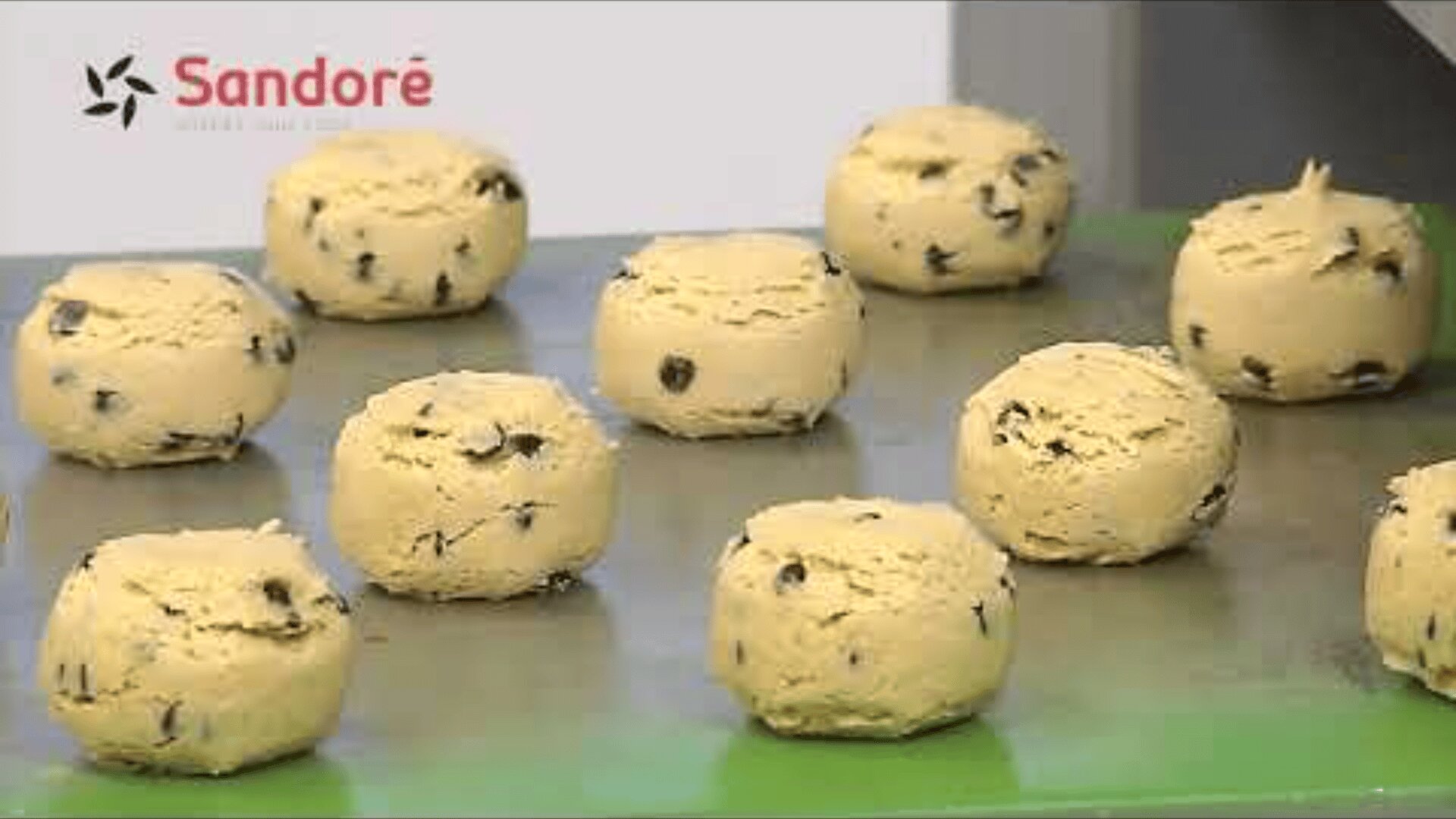 Mehrere runde Kugeln aus Schokoladenplätzchenteig sind auf einem Backblech angeordnet. Das Sandoré-Logo ist in der oberen linken Ecke des Bildes zu sehen.
