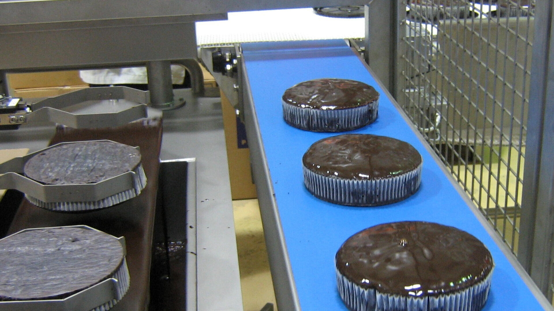 Schokoladenkuchen werden auf einer automatisierten Produktionslinie mit Schokoladenglasur überzogen, die auf einem blauen Förderband in einer Fabrikhalle läuft.