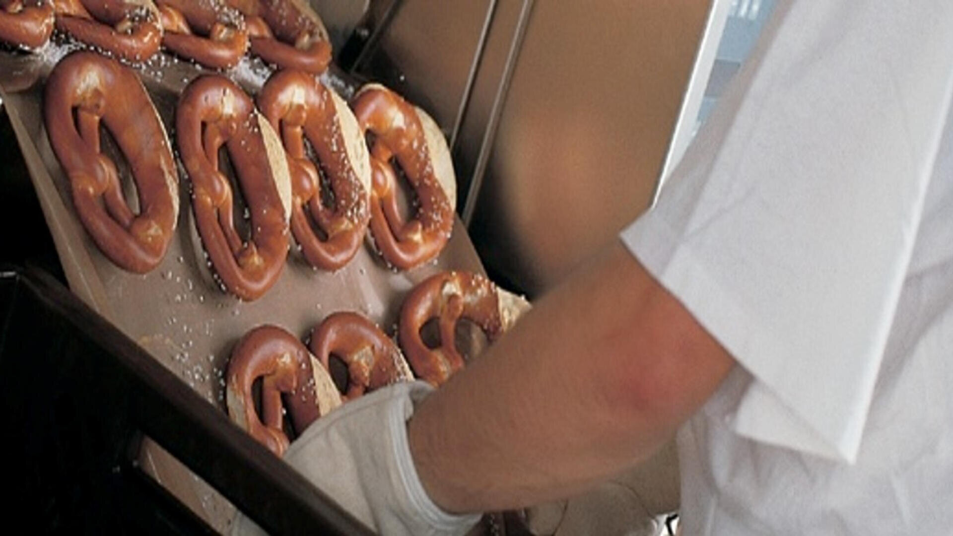 Eine Person mit weißem Hemd und Handschuhen holt ein Tablett mit frisch gebackenen, gesalzenen Brezeln aus einem Ofen.