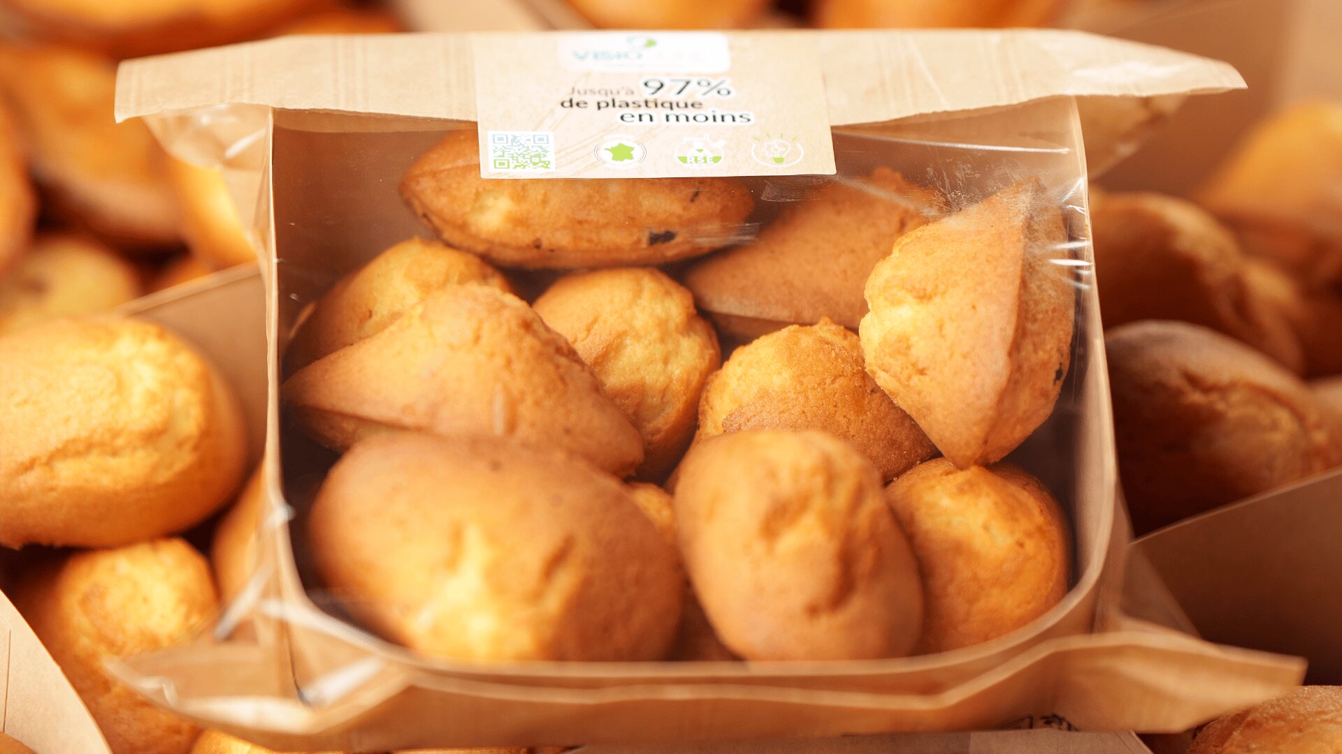 A close-up of a cardboard box filled with golden-brown madeleine pastries, partially covered by clear plastic. The box label mentions reduced plastic usage, with more pastries visible in the background.