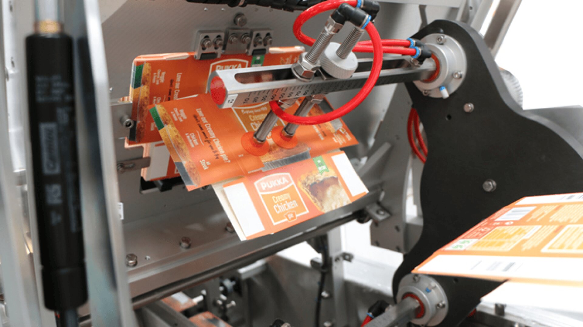 A packaging machine holding several orange Pukka chicken pie boxes with suction cups and mechanical arms, preparing to assemble or seal the packages.