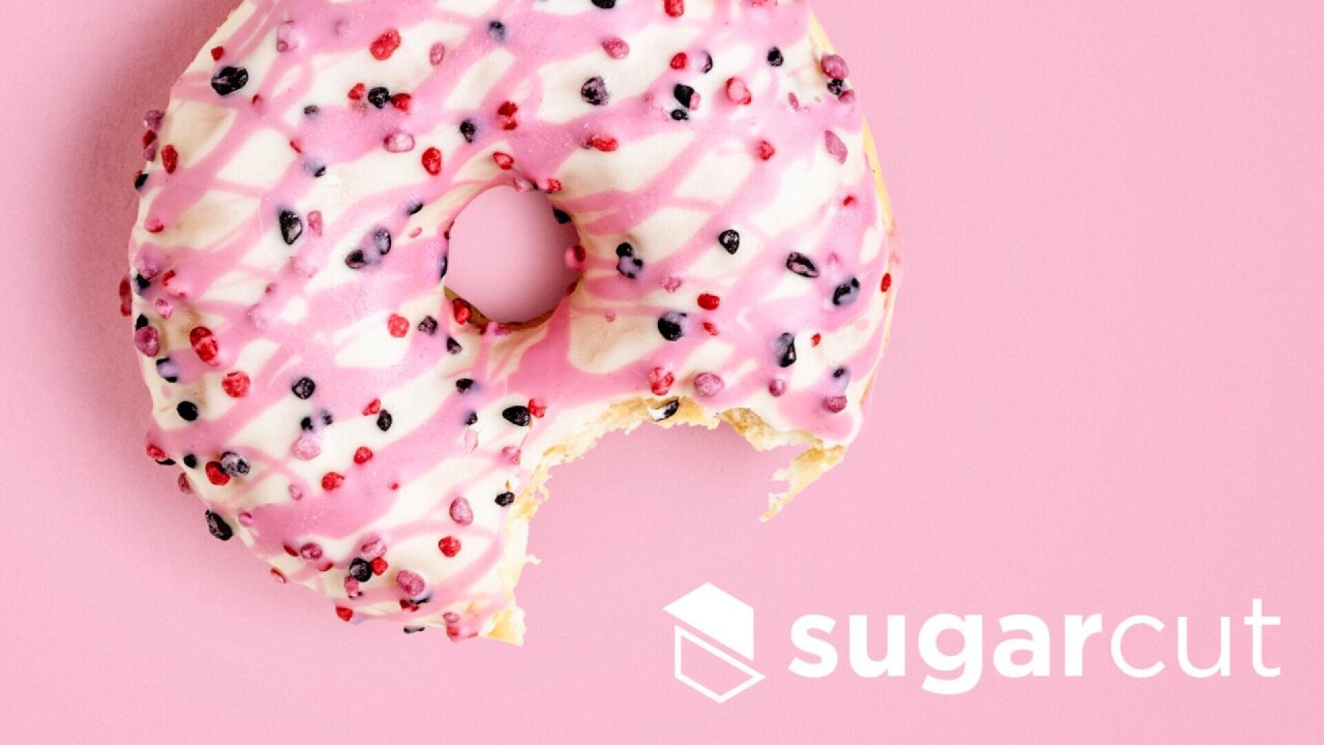 A donut with pink and white icing and colorful sprinkles, missing a bite, on a pink background. The word sugarcut and a cube-shaped logo appear in white at the bottom right.