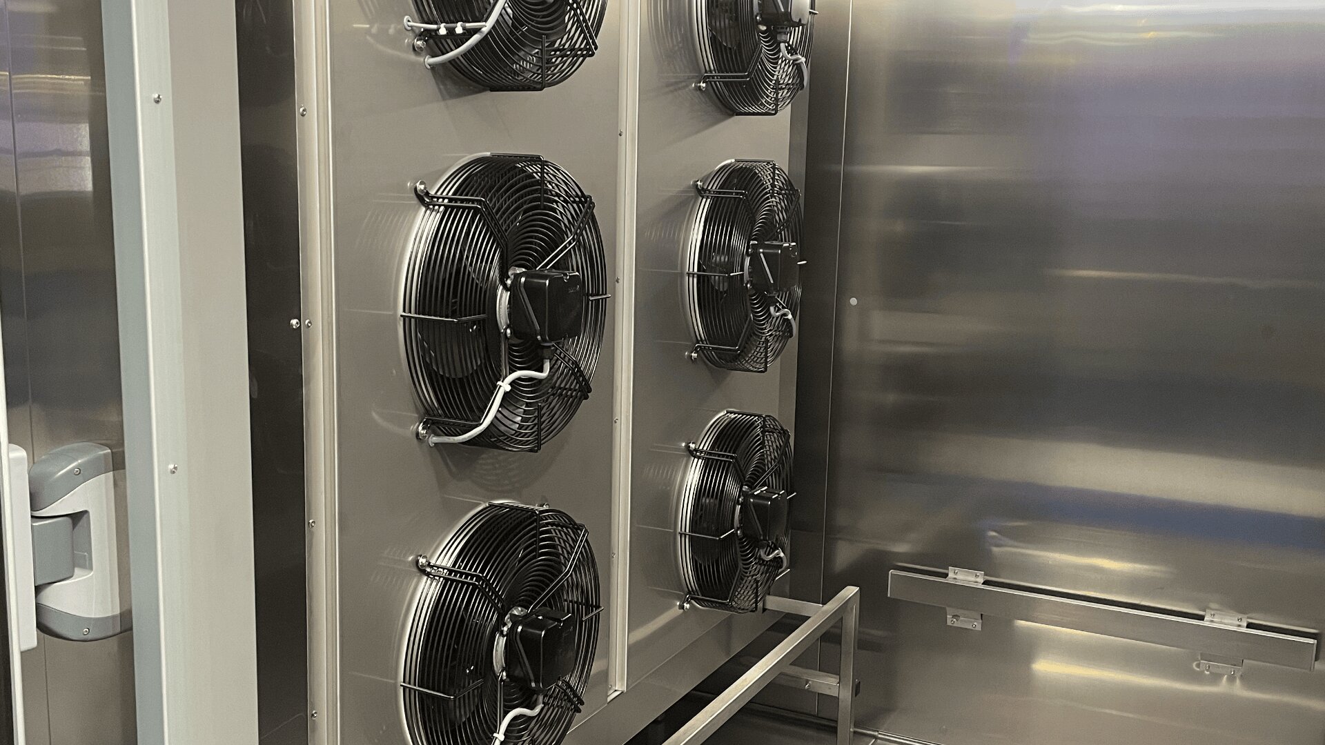 Four large black industrial fans are mounted vertically inside a metallic chamber, possibly used for ventilation or cooling purposes, with shiny metal walls and a metal frame structure in the foreground.