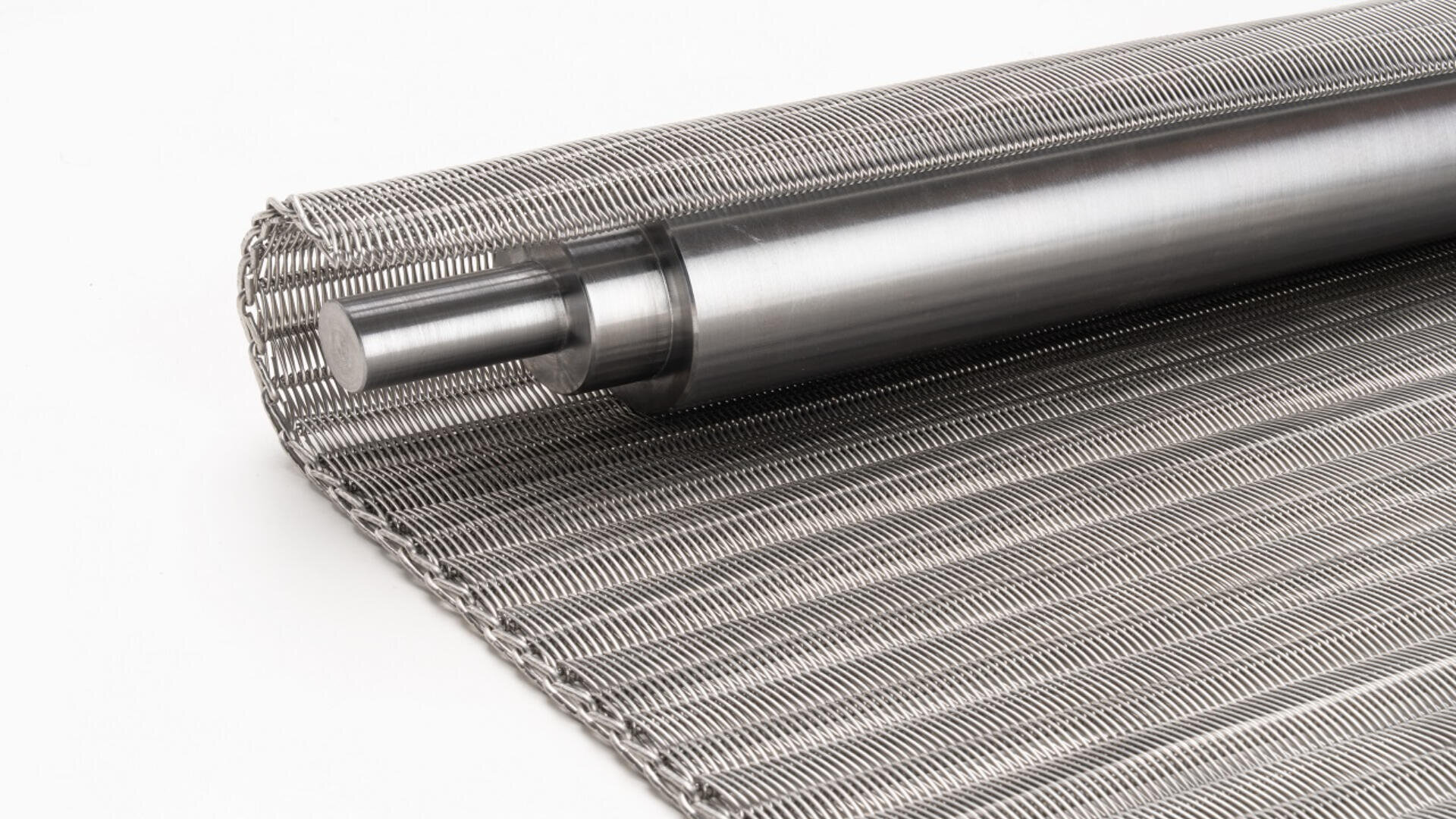 A close-up of a rolled-up metal conveyor belt with interwoven wires and a cylindrical metal shaft, placed on a white background.