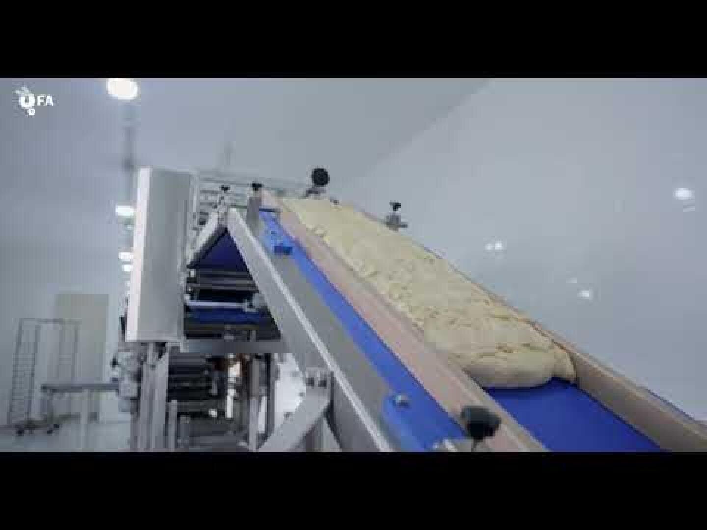 A large piece of dough moves along a blue conveyor belt in a clean, brightly lit industrial food processing facility.
