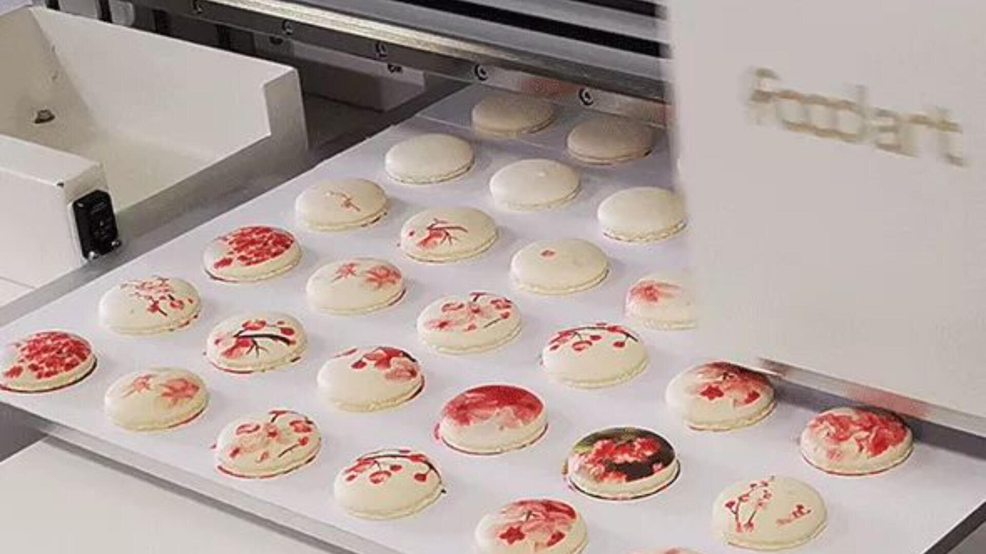 A machine is printing red and pink floral designs onto white macarons arranged in rows on a tray.