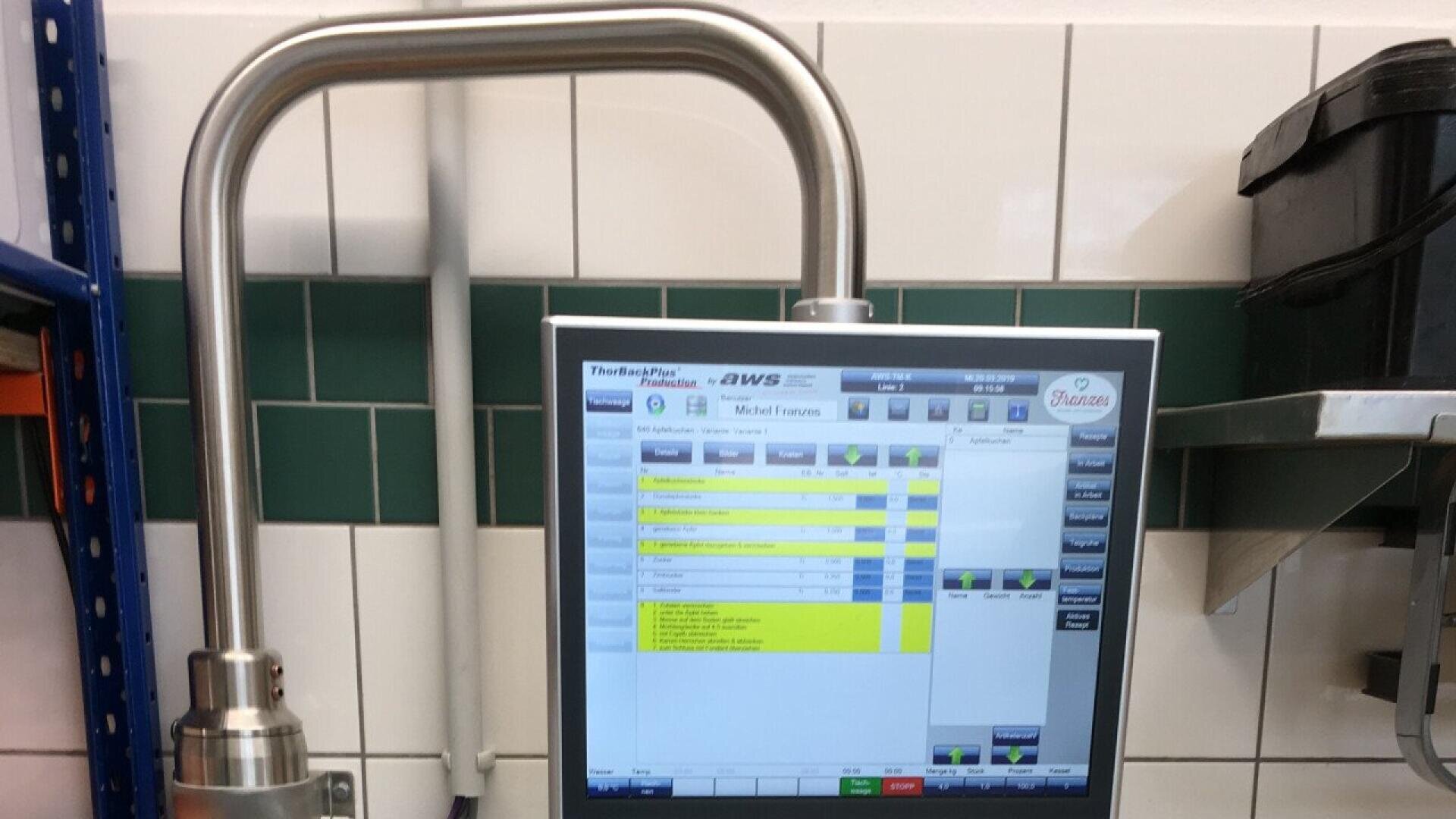 A close-up of a touchscreen control panel displaying a software interface in a tiled industrial or laboratory setting, with green and white tiles and metal equipment visible in the background.