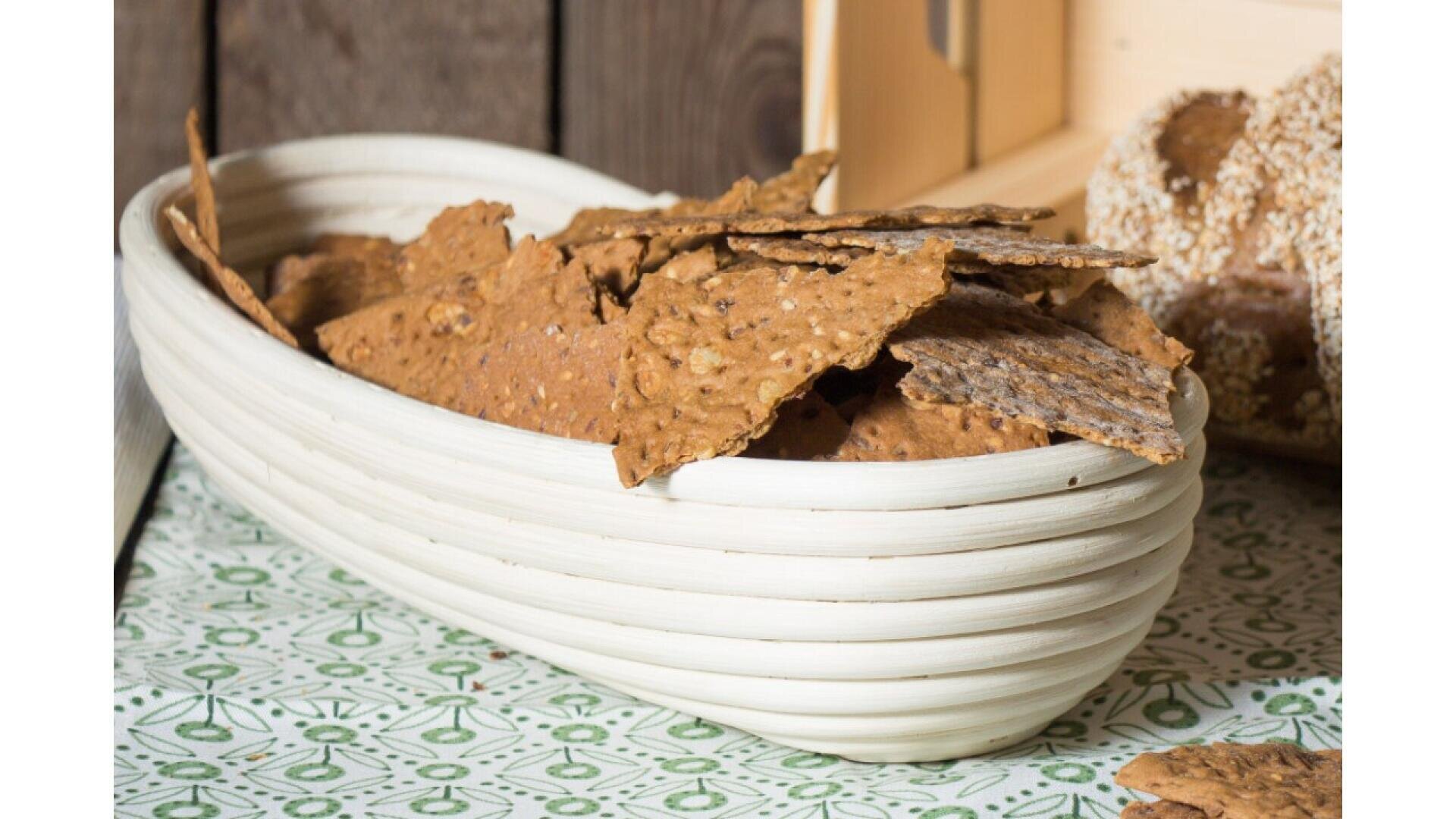 Ein weißer, ovaler Korb enthält mehrere knusprige Körnercracker, die auf einem grün-weiß gemusterten Tuch liegen, mit Brot im Hintergrund.