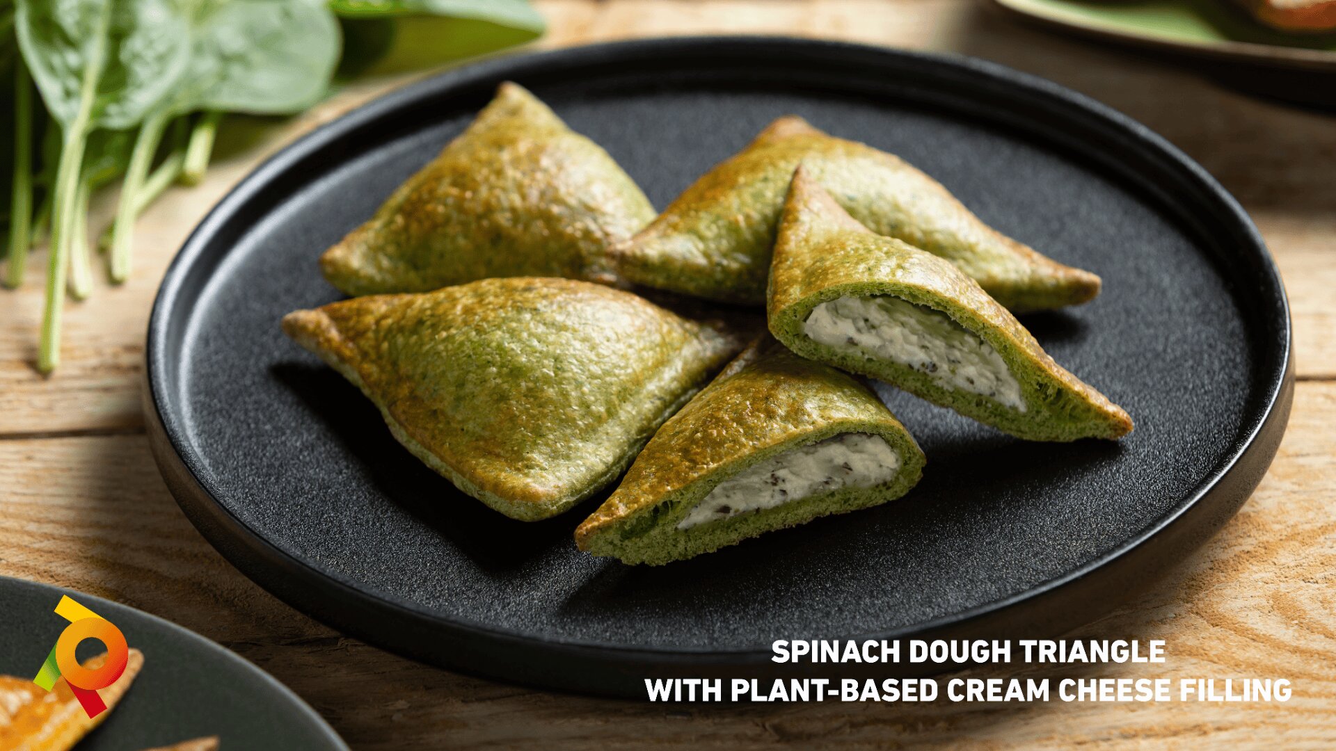 Four spinach dough triangles filled with plant-based cream cheese are arranged on a black plate, with fresh spinach leaves in the background. Text at the bottom reads: “Spinach dough triangle with plant-based cream cheese filling.”.