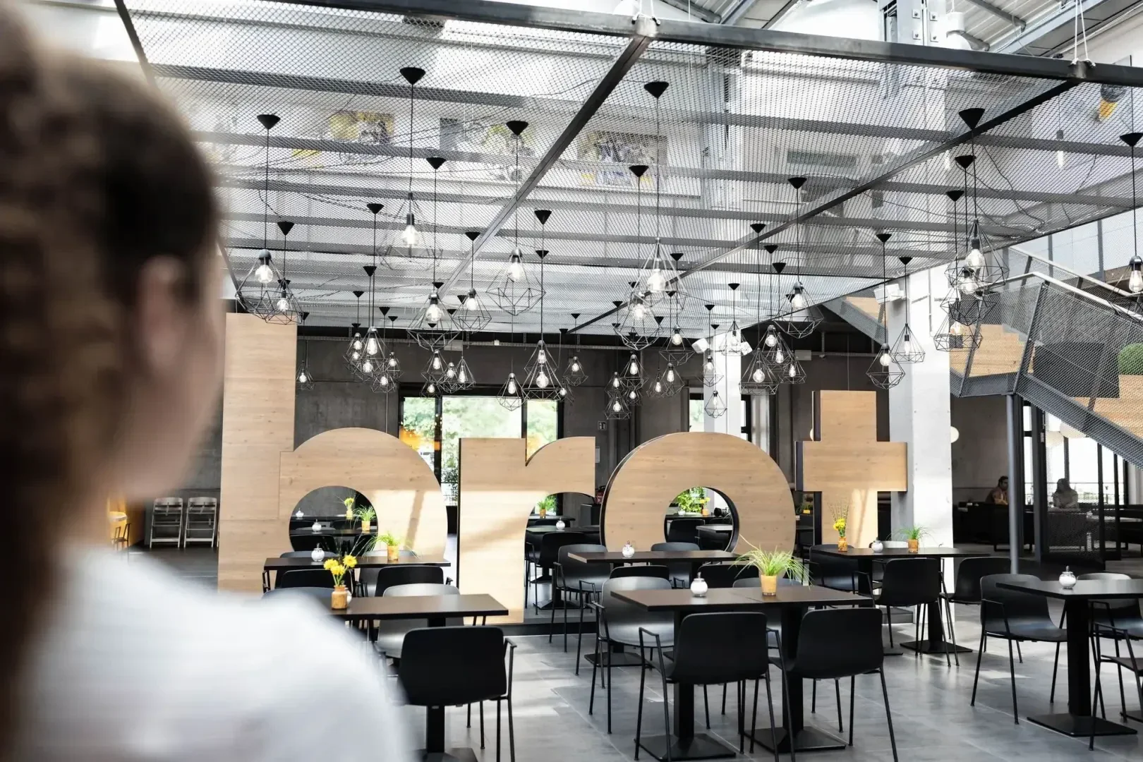 Eine moderne, geräumige Cafeteria mit schwarzen Tischen und Stühlen, Hängelampen und großen Holzbuchstaben mit der Aufschrift brot im Hintergrund. Eine Person mit lockigem Haar ist unscharf im Vordergrund zu sehen.