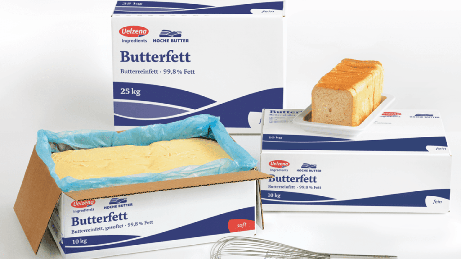 Boxes labeled Butterfett (butterfat) in German, with one open box showing a large slab of butter, a loaf of bread on a box, and a metal whisk in the foreground.