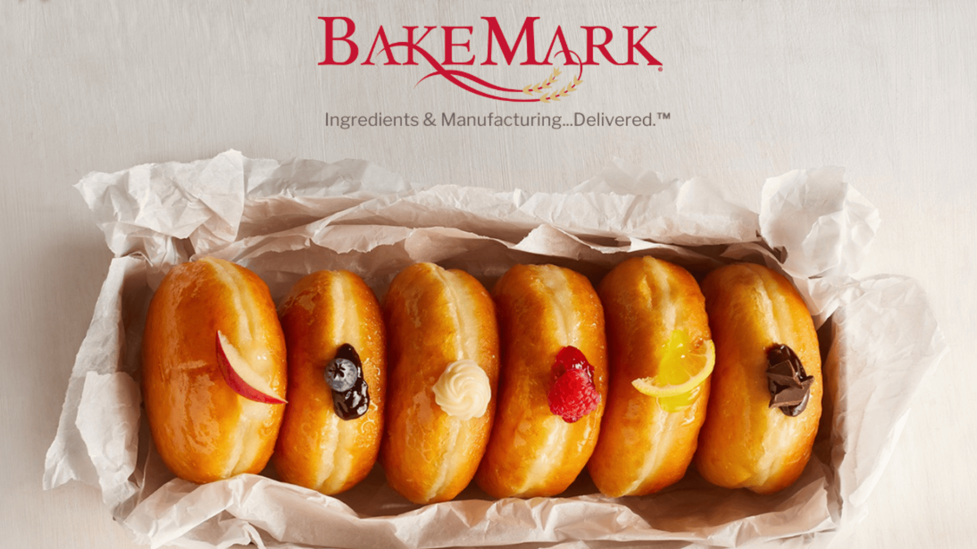Six glazed donuts with various fillings and toppings are arranged in a row on parchment paper. Above them is the BakeMark logo with the tagline, “Ingredients & Manufacturing...Delivered.”.